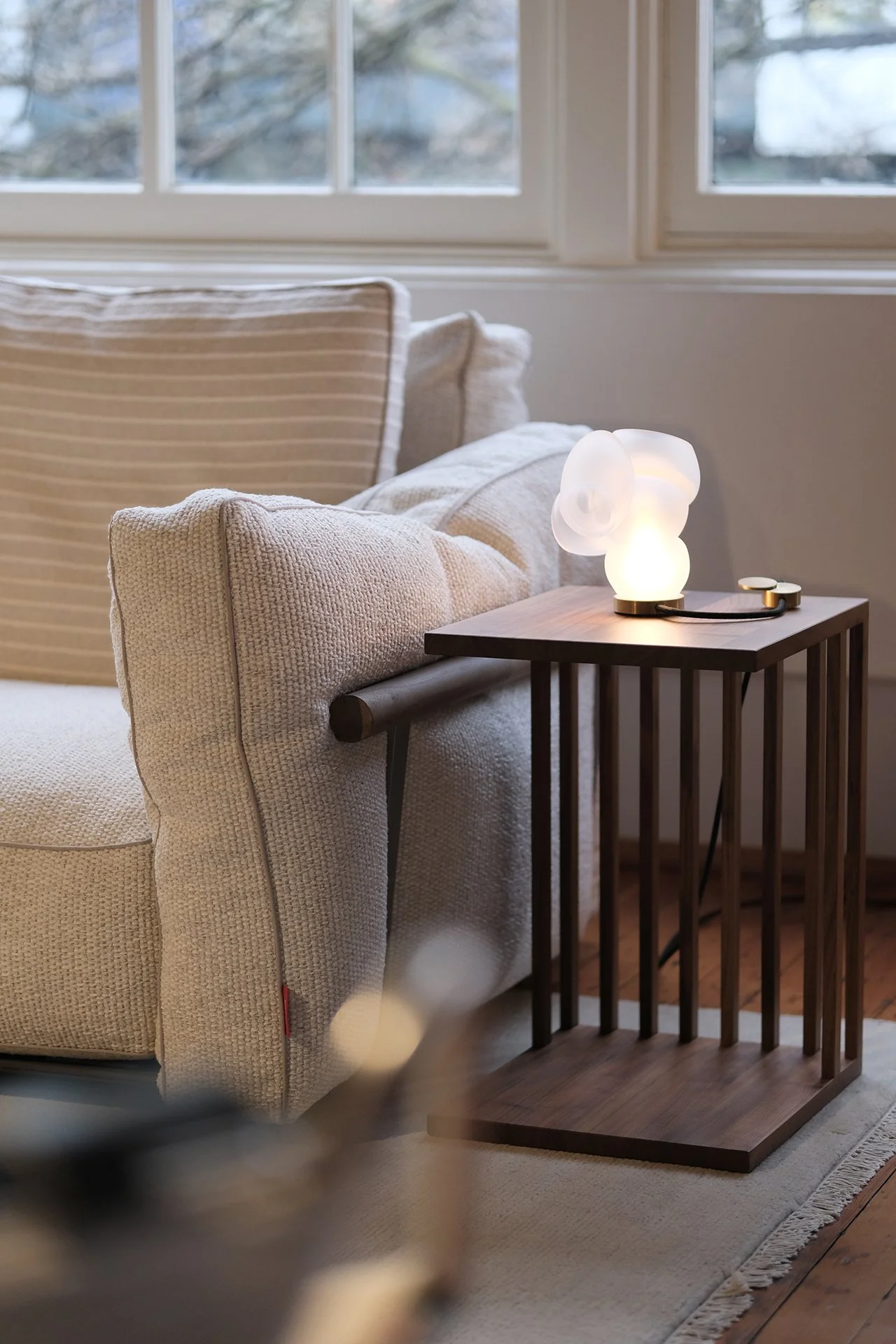 A cozy living room corner with a beige textured sofa, a wooden side table, and a modern lamp with a sculptural design, near a window showing trees outside.