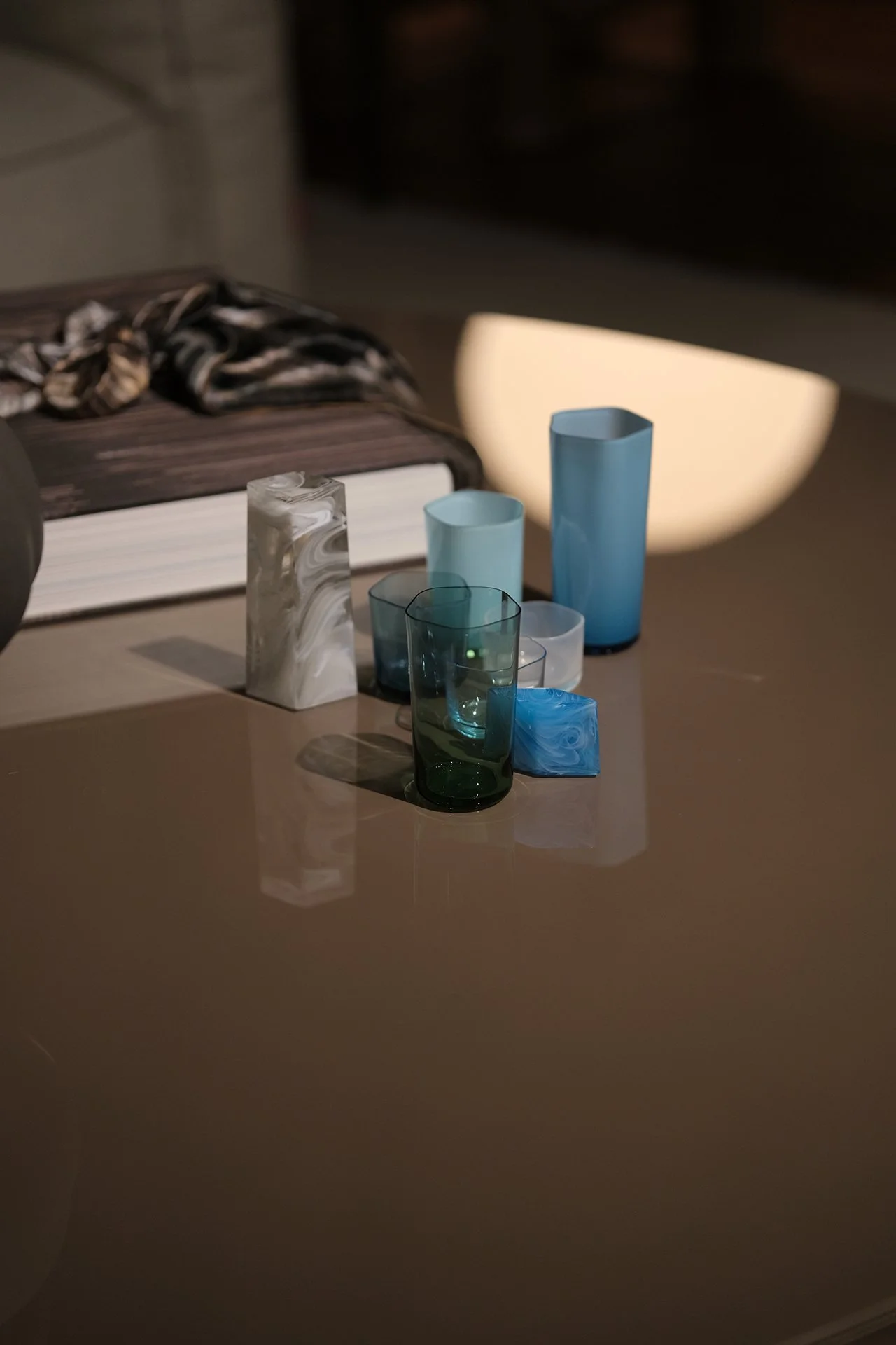 An assortment of colorful glass vases and bowls on a reflective surface, with a large book and a rounded lit object in the background.