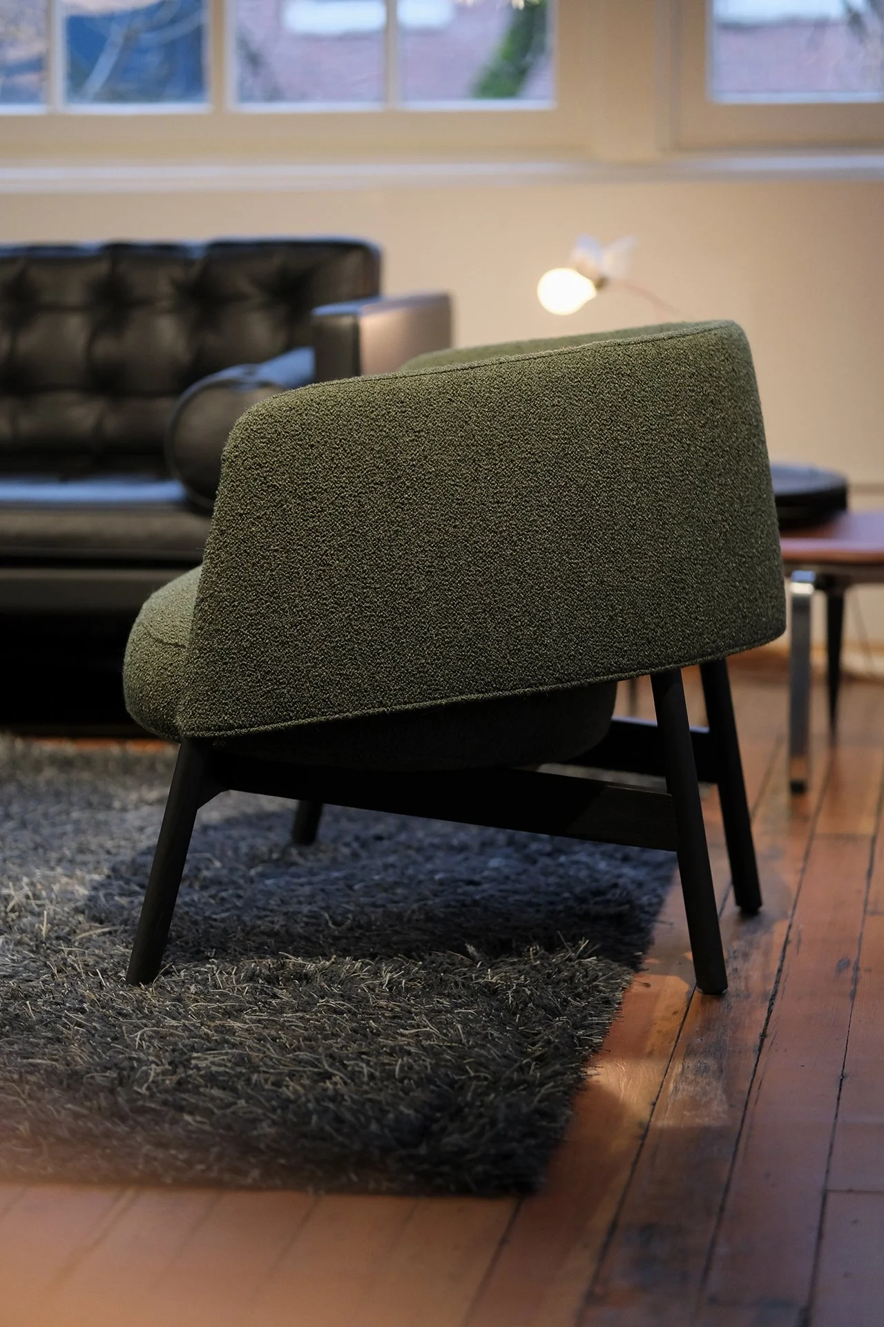 A green upholstered armchair with black wooden legs in a living room with wooden floors, a black textured rug, and a black tufted leather sofa in the background near a window.