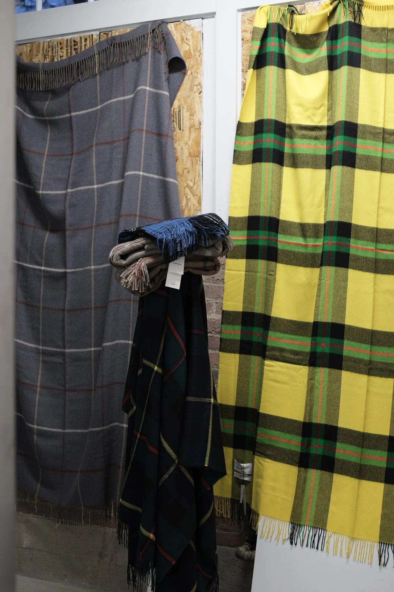 Display of plaid blankets and throws in various colors and patterns, including gray, yellow, and dark plaid, hung on a wall and draped over a stand.