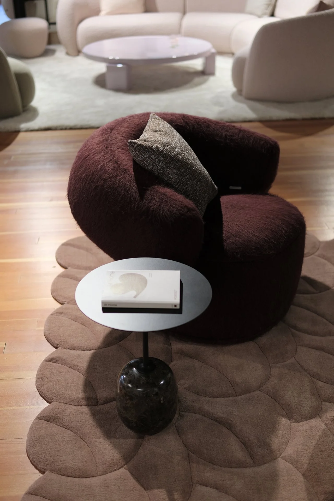 A cozy lounge area featuring a plush, brown, circular chair with a textured cushion, a small side table with a book, and background of white and beige sofas with a circular coffee table.
