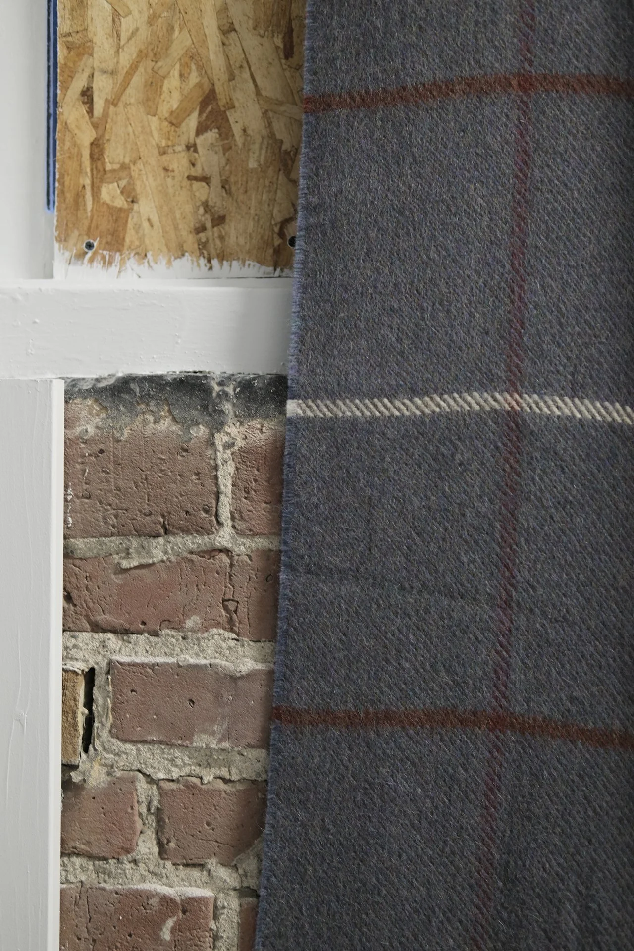 Close-up of a corner showing a brick wall, a white painted wooden trim, an OSB wall panel, and a gray carpeted floor with red and white plaid pattern.