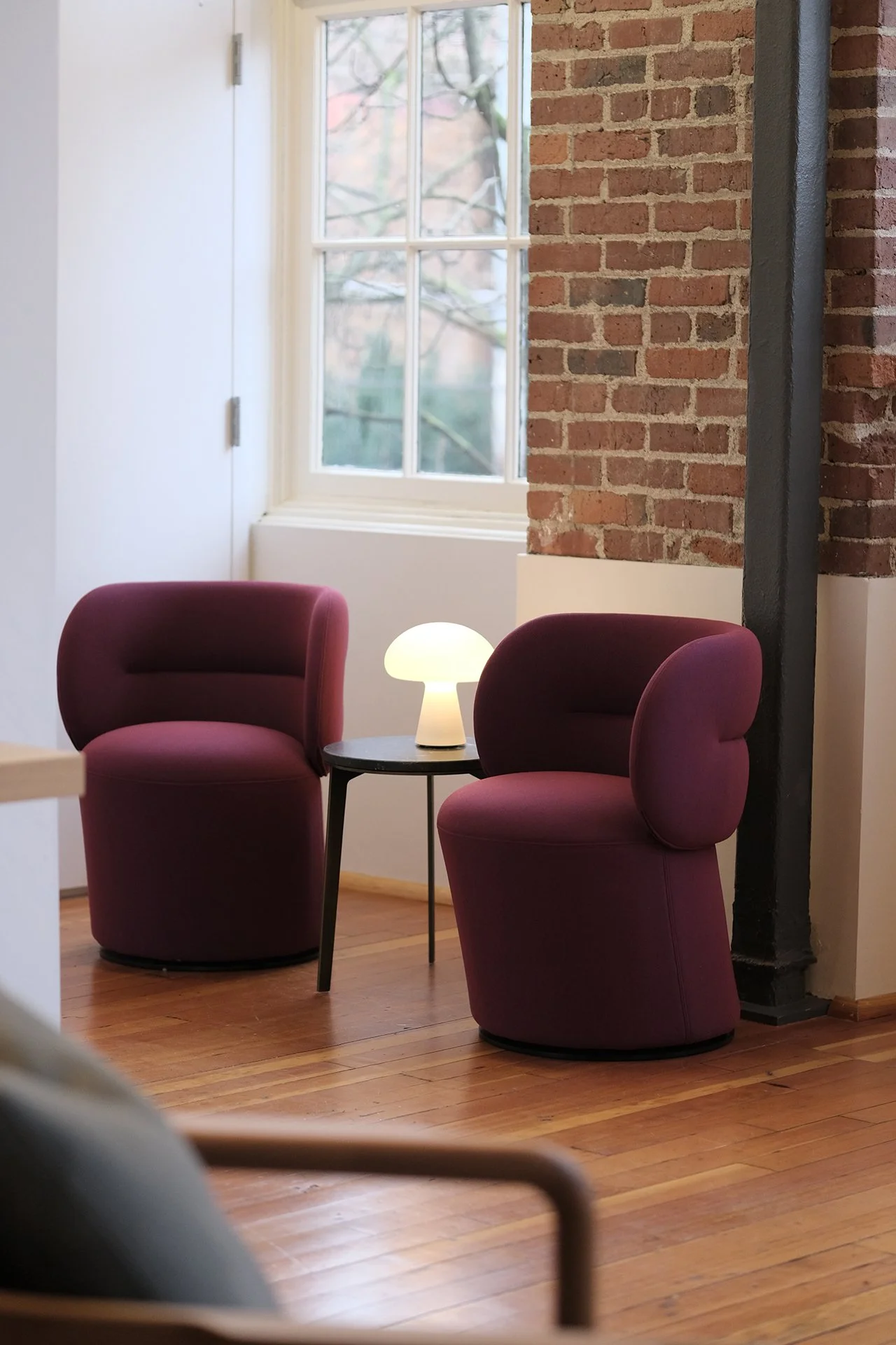 Two burgundy armchairs with rounded backs and a small black table holding a white lamp between them, in a room with a large window, exposed brick wall, white walls, and hardwood floors.