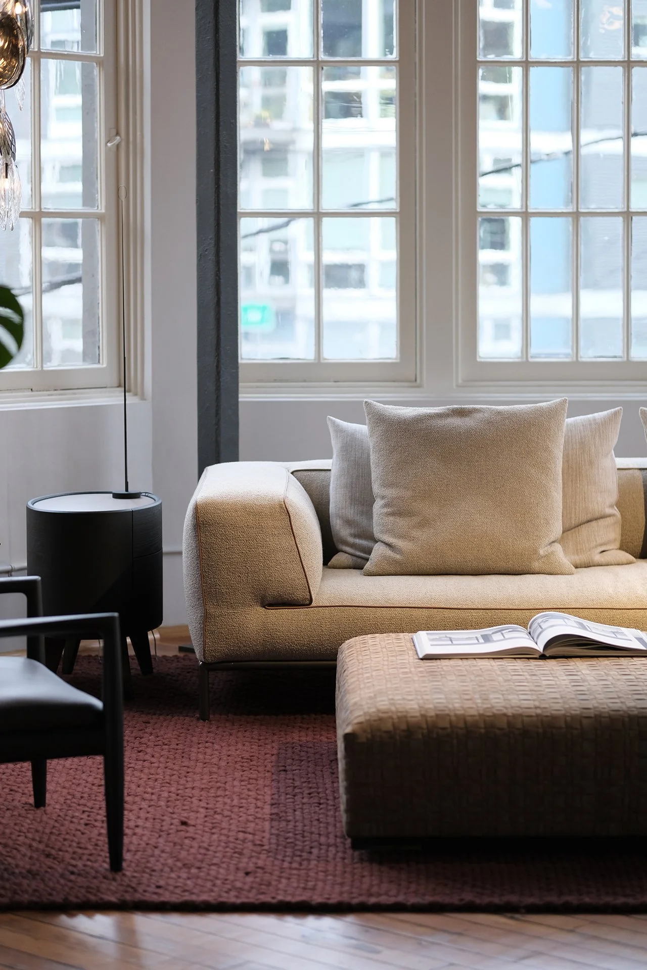A cozy living room with large windows, a beige sofa with matching pillows, a textured ottoman with an open book on it, a red rug, a black side table with a lamp, and a black chair.