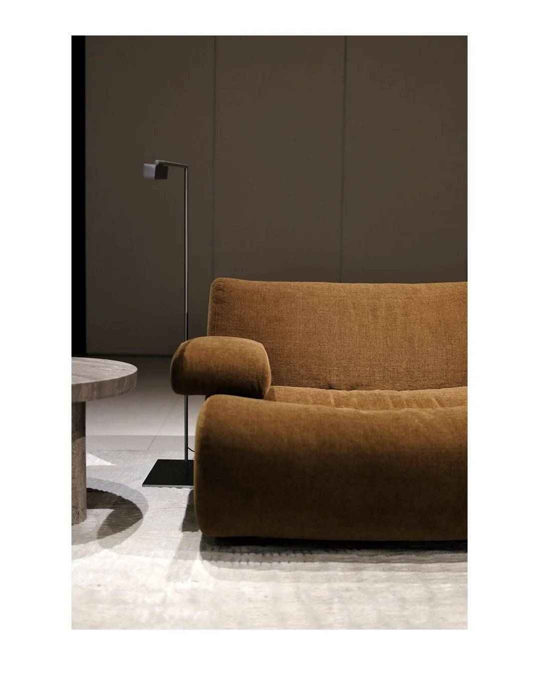 Interior of a modern living room featuring a brown fabric sofa, a small black side table, a tall black floor lamp, and a beige rug with a light-colored floor and dark wall in the background.