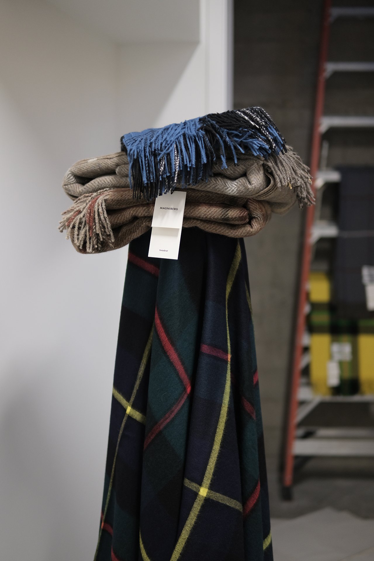 Folded scarves on a stand with a store tag, featuring a plain beige, a beige with red lines, and a dark green plaid pattern scarf.