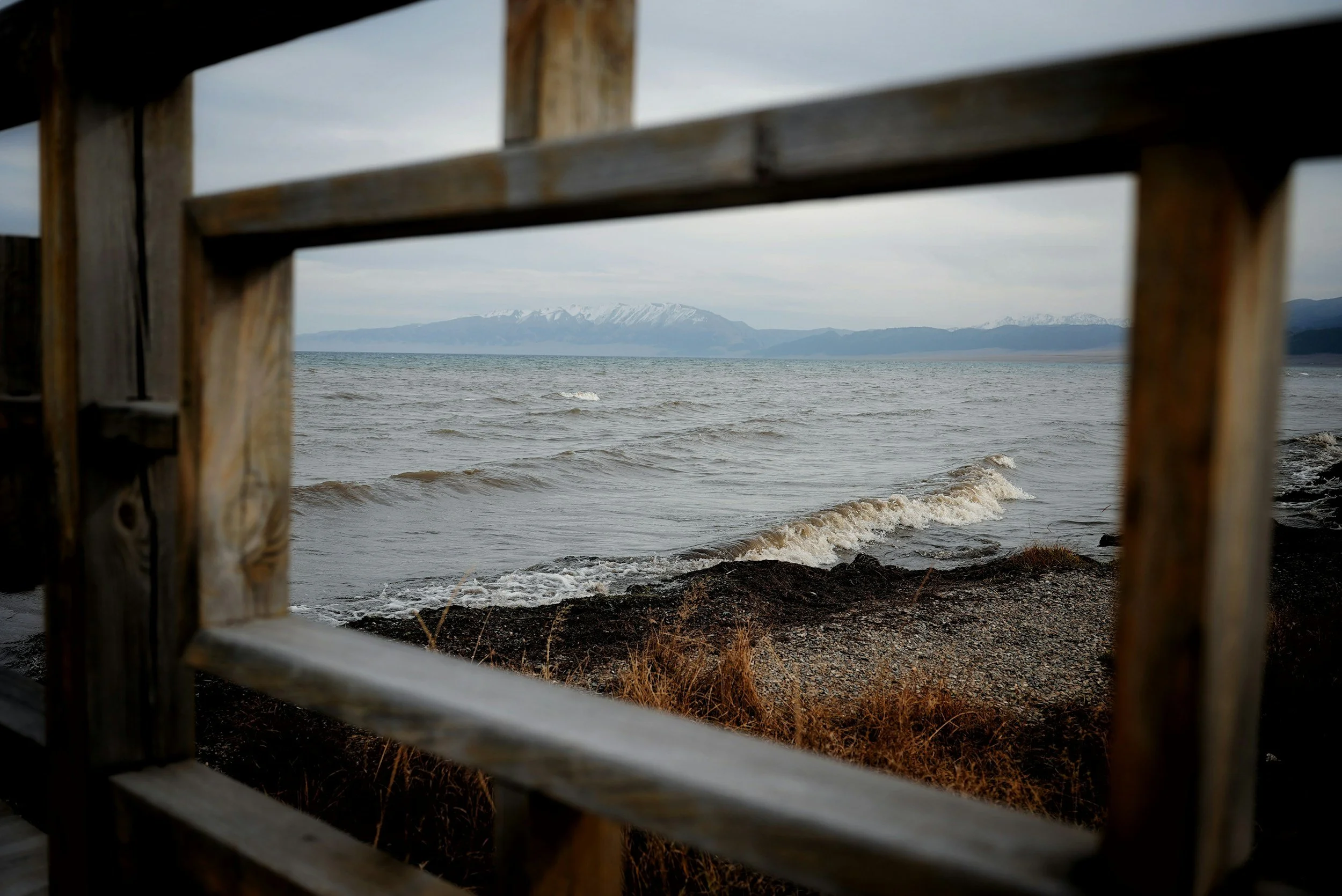 A view of ocean waves and a pebble shoreline framed by a wooden window, representing a contained act of poetic witnessing.