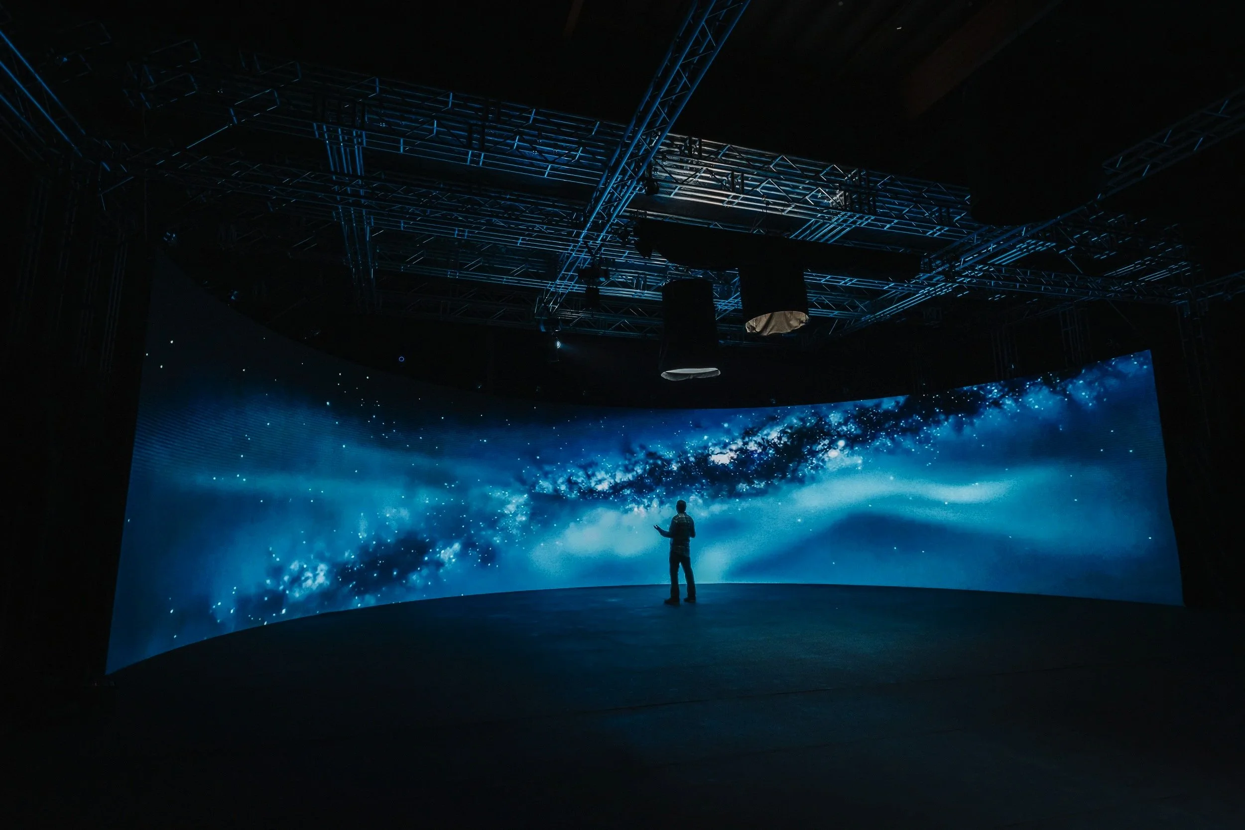 Person standing in front of a large curved screen displaying a starry galaxy or outer space scene with blue and white colors, in a dark room with industrial lighting structure overhead.