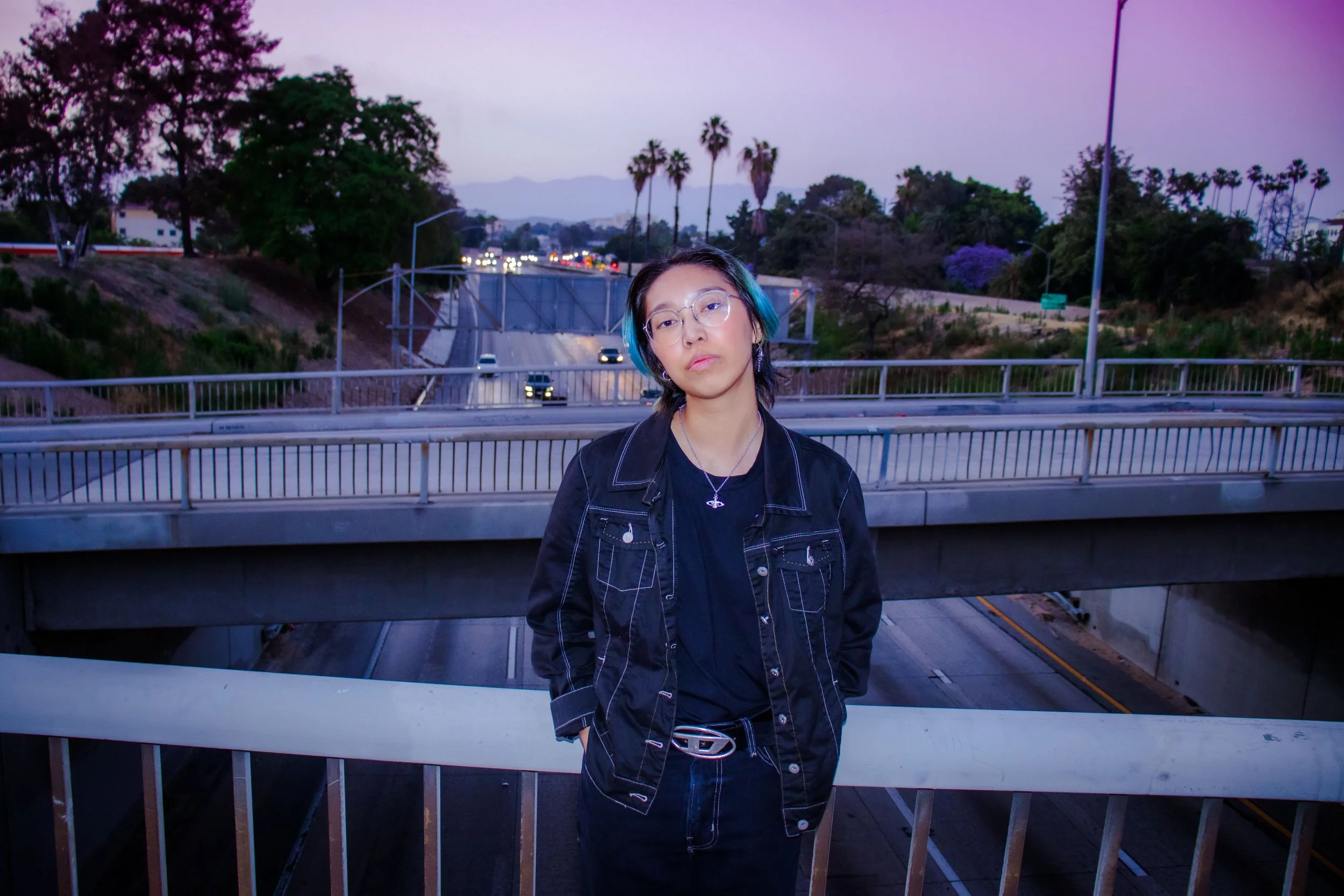 A young woman with short black hair, glasses, and blue highlights, standing on a bridge over a highway at dusk, wearing a black jacket and black clothing, with trees and palm trees in the background.
