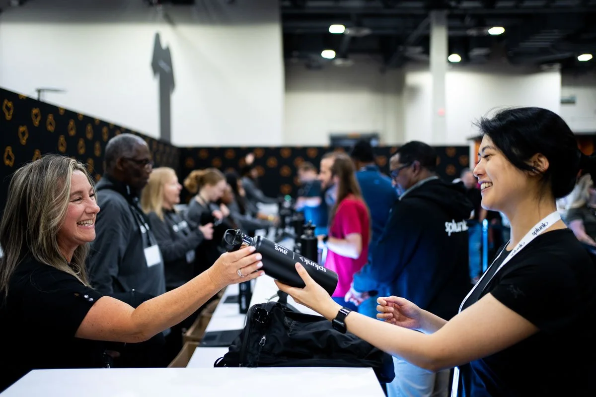Two women at a conference or trade show exchanging a water bottle. The woman on the left is smiling and has light brown hair, while the woman on the right is also smiling and has dark hair. There are several people in the background attending the event.