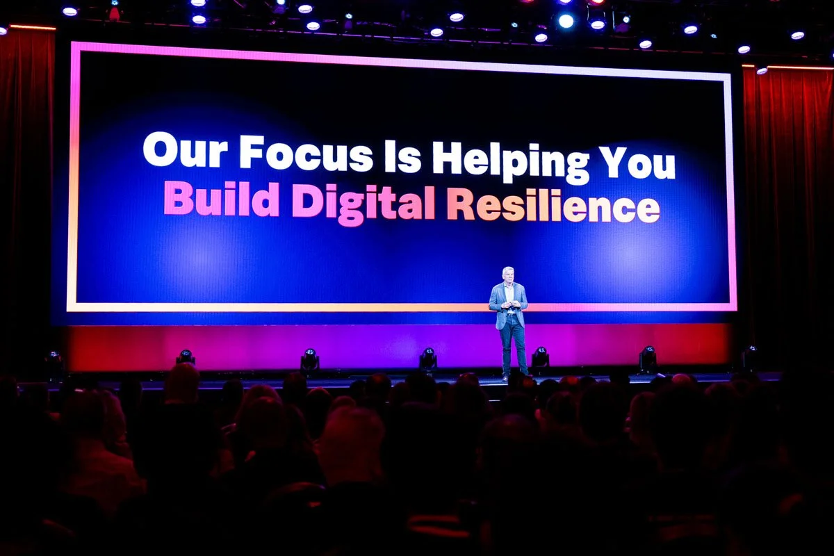 A speaker on stage at a conference with a large screen behind him displaying the message, 'Our Focus Is Helping You Build Digital Resilience', with an audience seated in front.