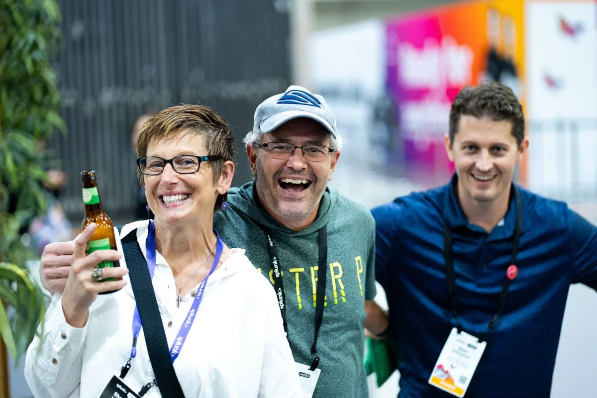 Three cheerful adults at a conference or event, smiling and standing close together. The woman on the left holds a beer bottle, the man in the middle wears glasses and a cap, and the man on the right wears a blue shirt. All have conference badges around their necks.
