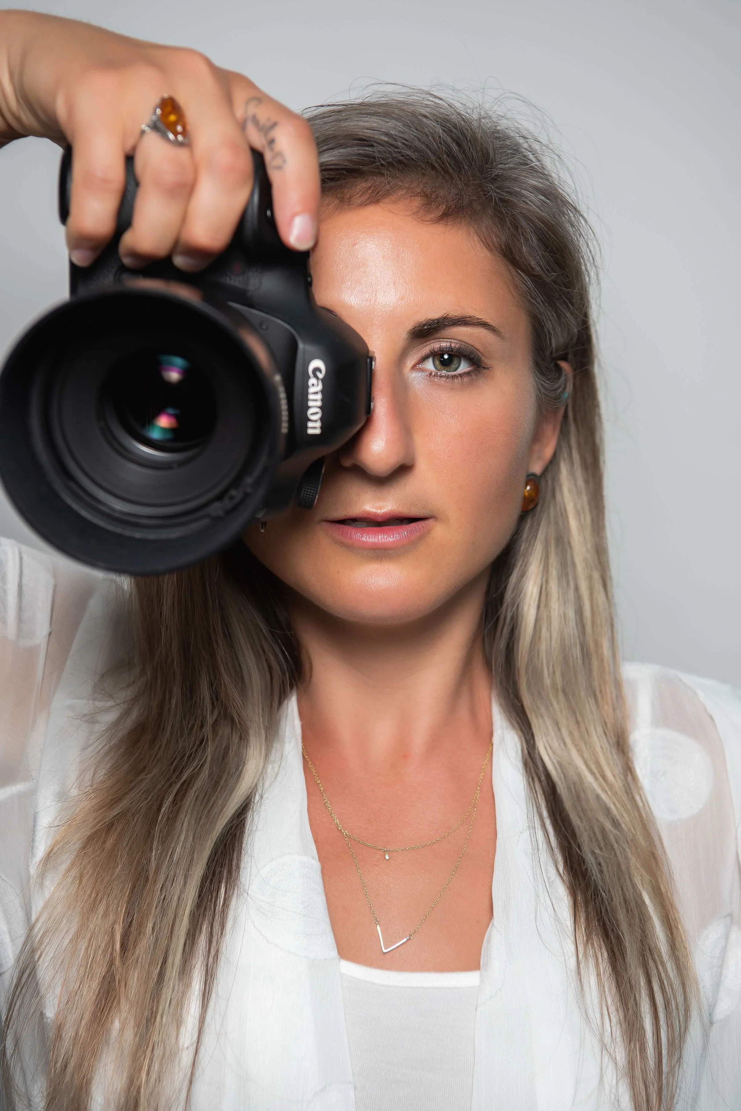 A woman with long, wavy hair and earrings is taking a selfie with a DSLR camera, covering her left eye. She wears a white top and layered gold necklaces.