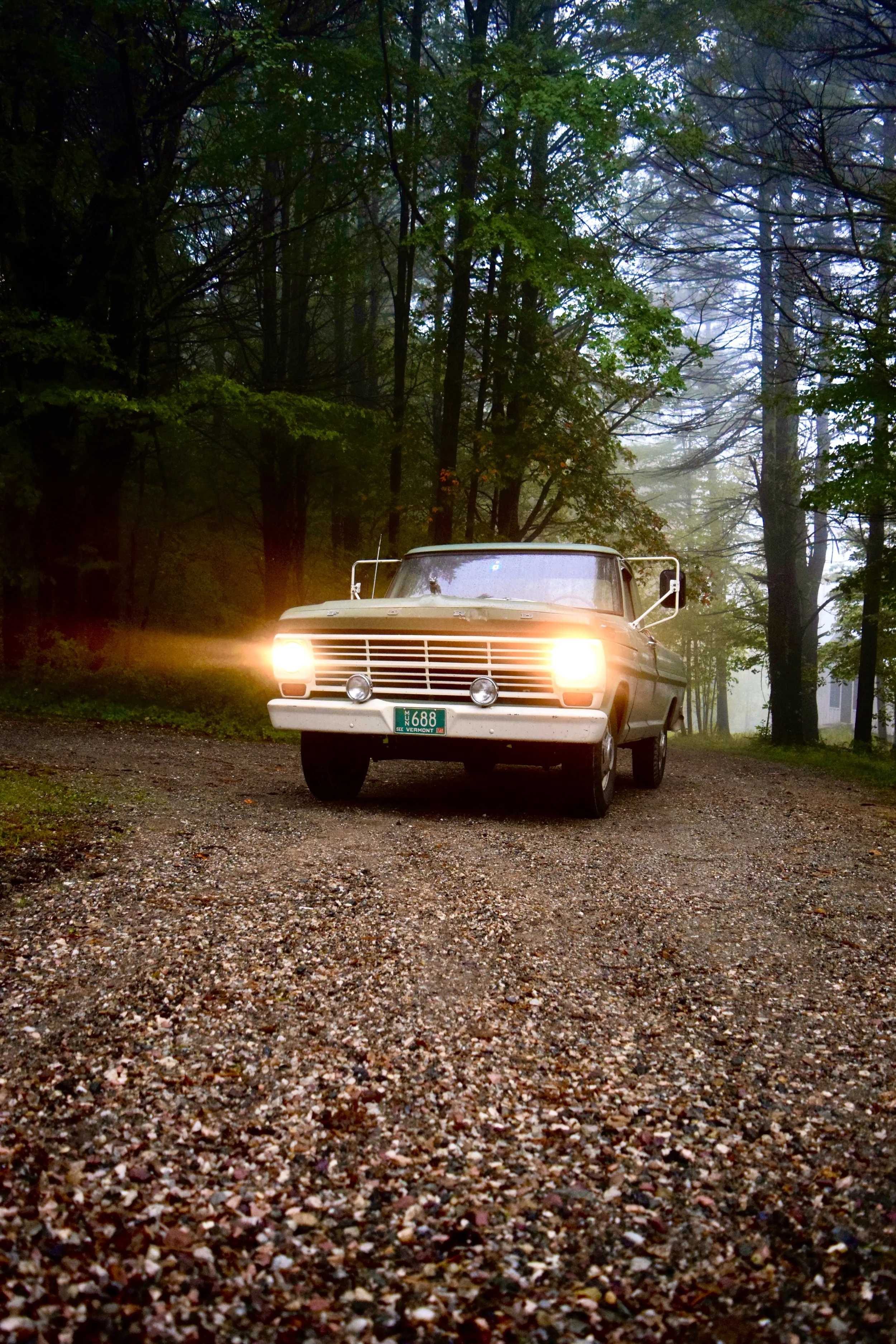 A truck with blazing headlights drives up a forest drive in the rain.