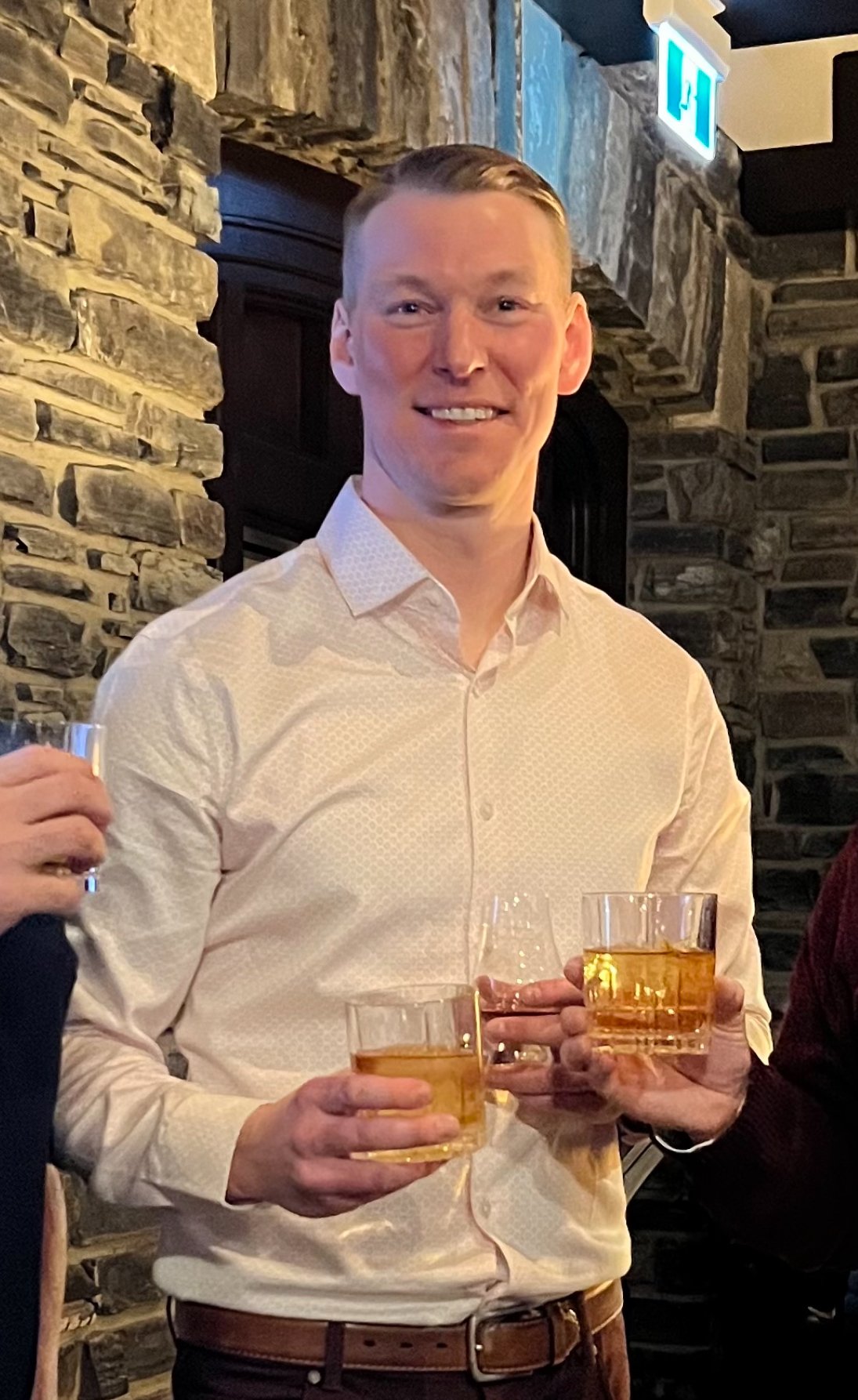 A man in a cream-colored dress shirt holding a glass of whiskey, smiling, at a social gathering in a restaurant or bar with stone walls and an exit sign overhead.