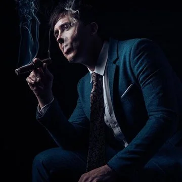 A man in a suit smoking a cigar with smoke rising in a dark setting.