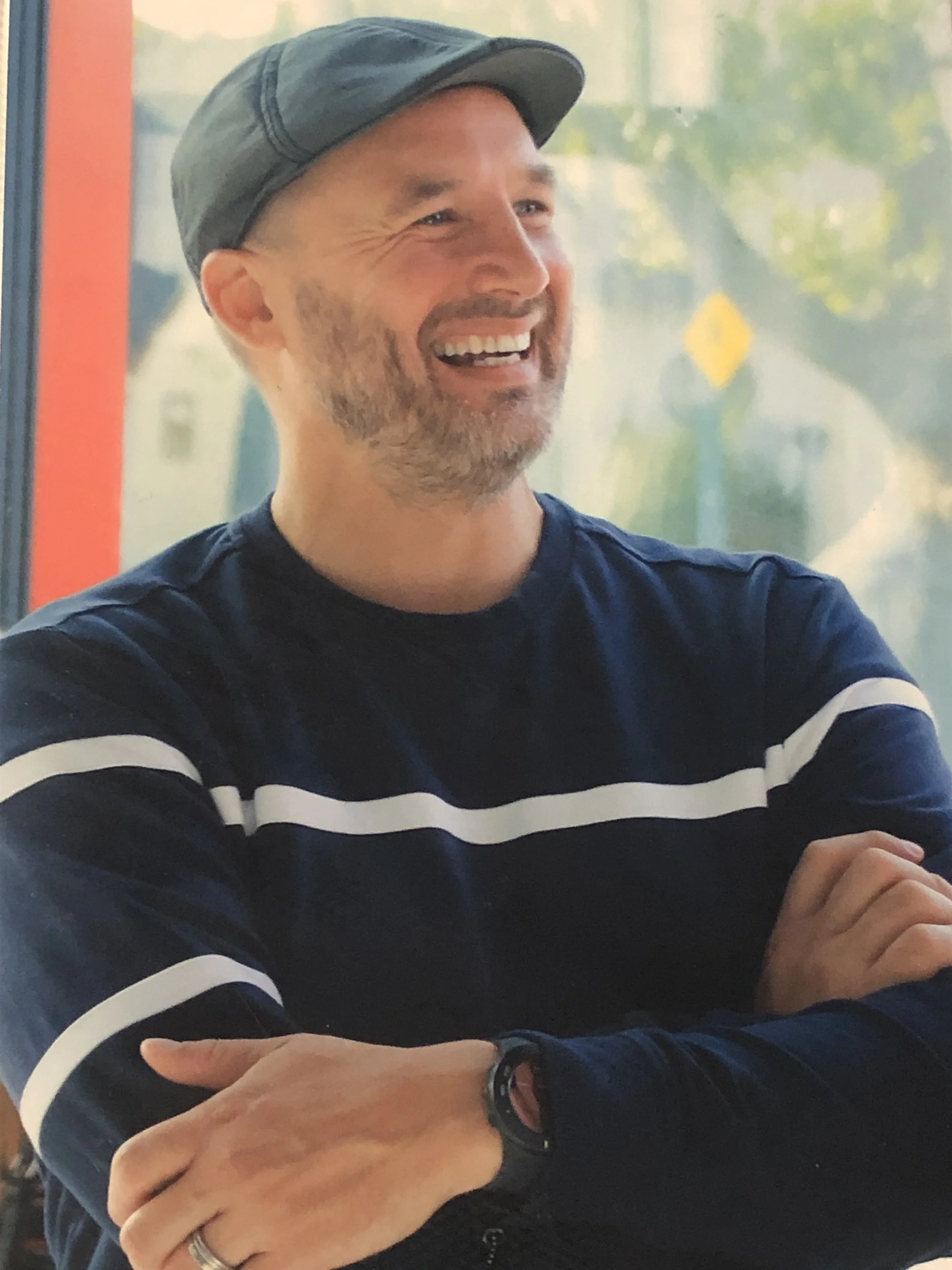 A smiling man wearing a dark hat and navy shirt with white stripes, sitting with his arms crossed and looking to the side.