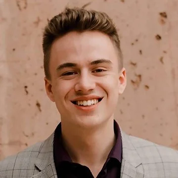 Young man smiling, wearing a gray checkered blazer and dark shirt, standing in front of a tan background.