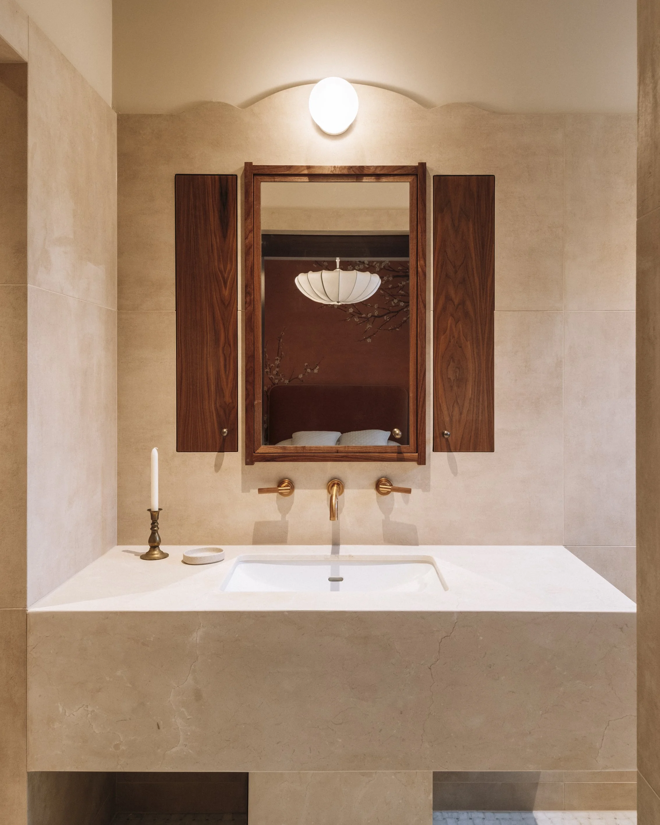 Modern bathroom with beige stone vanity, wall-mounted sink and mirror, brass faucet, candle holder, and wall light above.