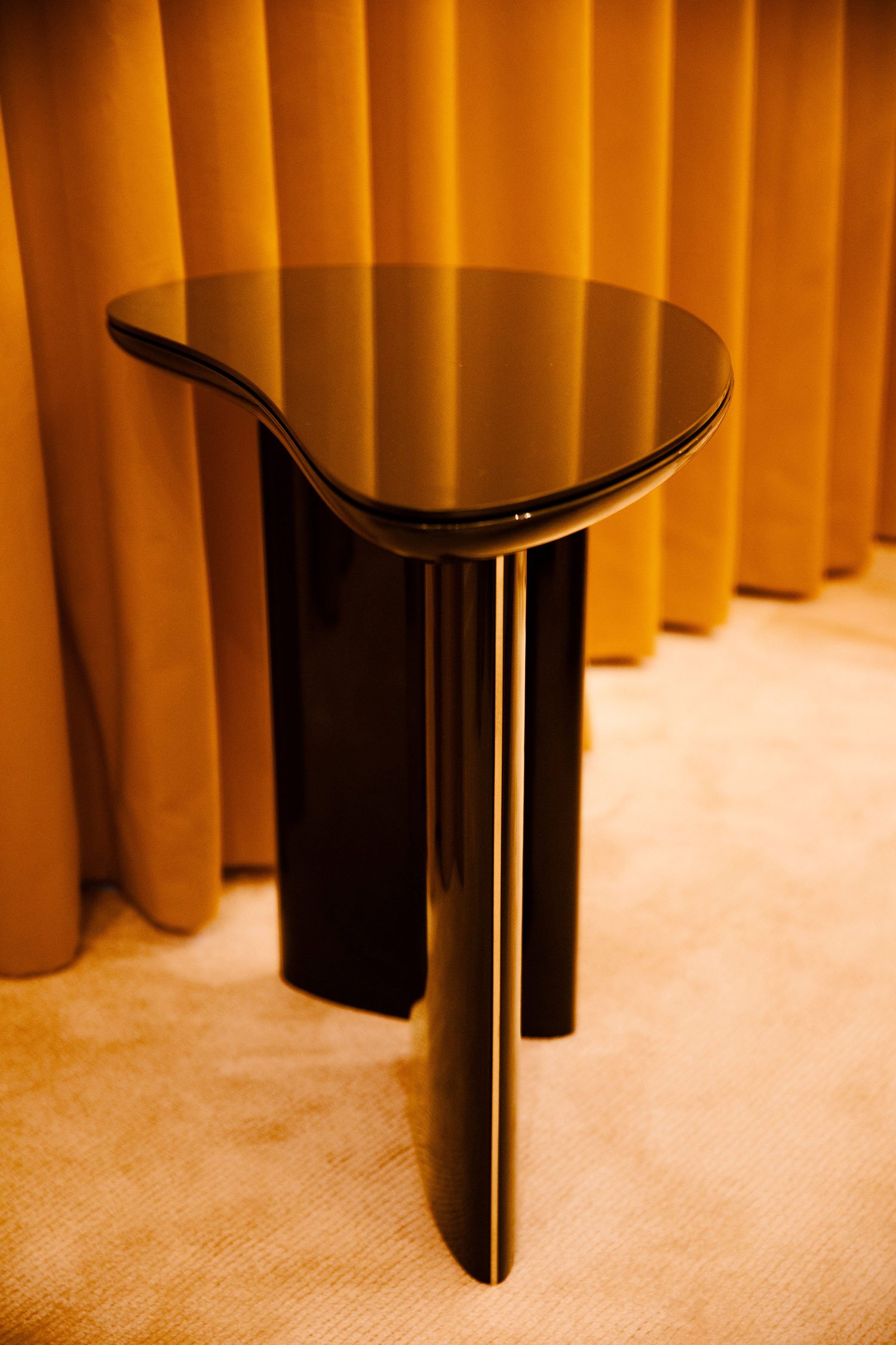 Modern black side table with an irregular shape, set against a beige curtain background.