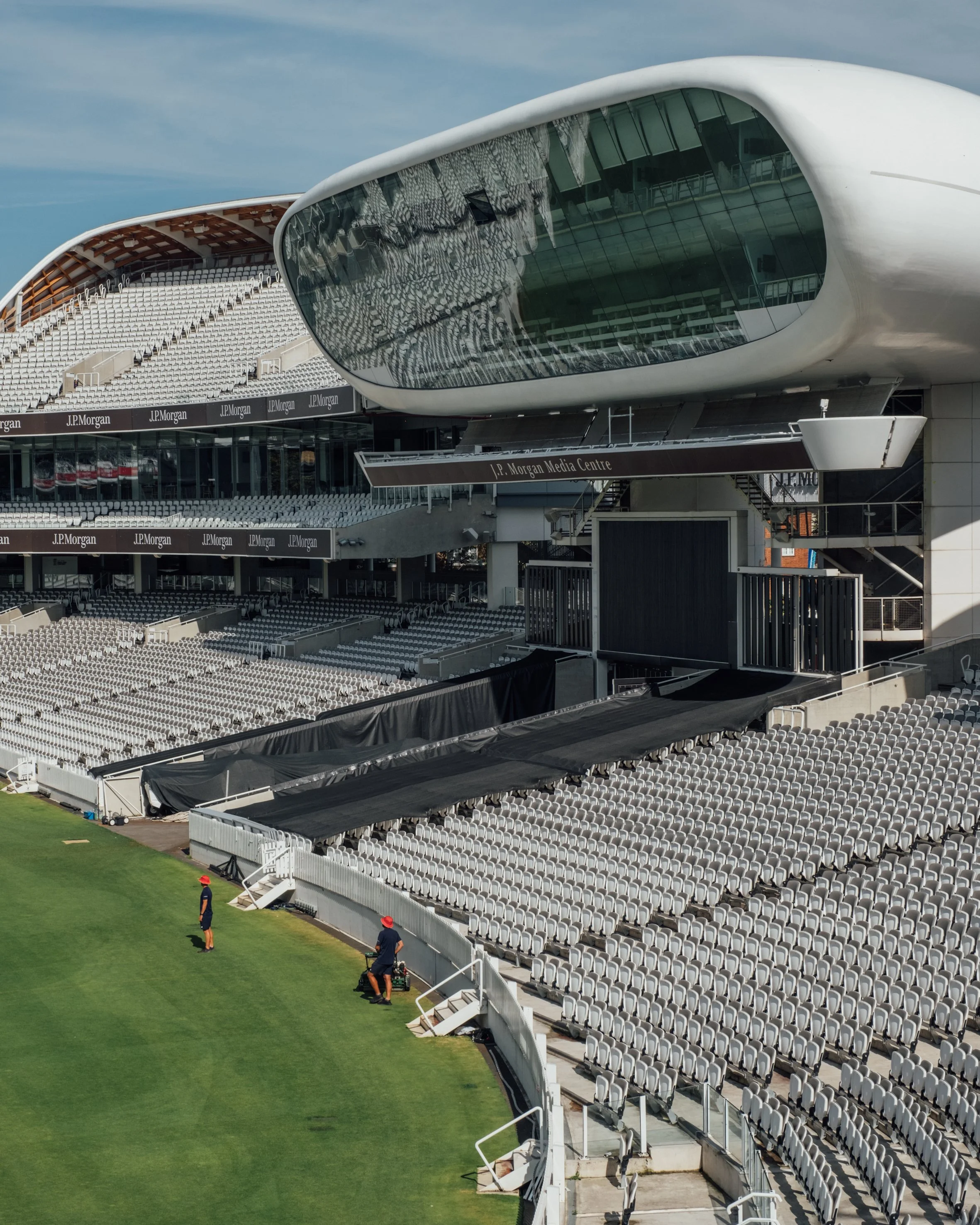 Empty sports stadium with large media center, rows of seating, and workers maintaining the field.