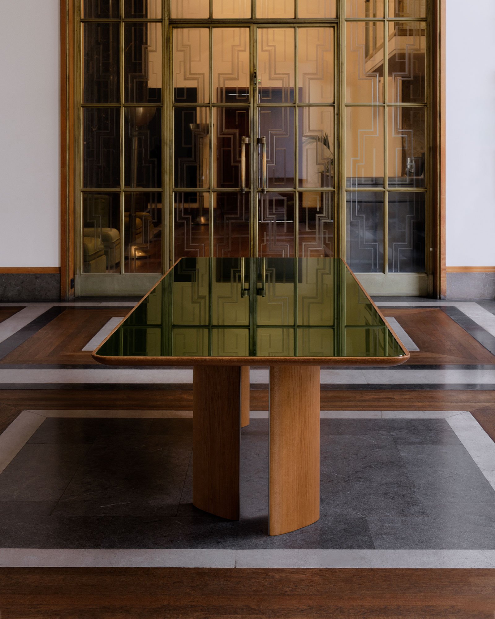 A modern table with a green-tinted glass top and wooden base in a sophisticated interior setting featuring a patterned floor and decorative glass doors.
