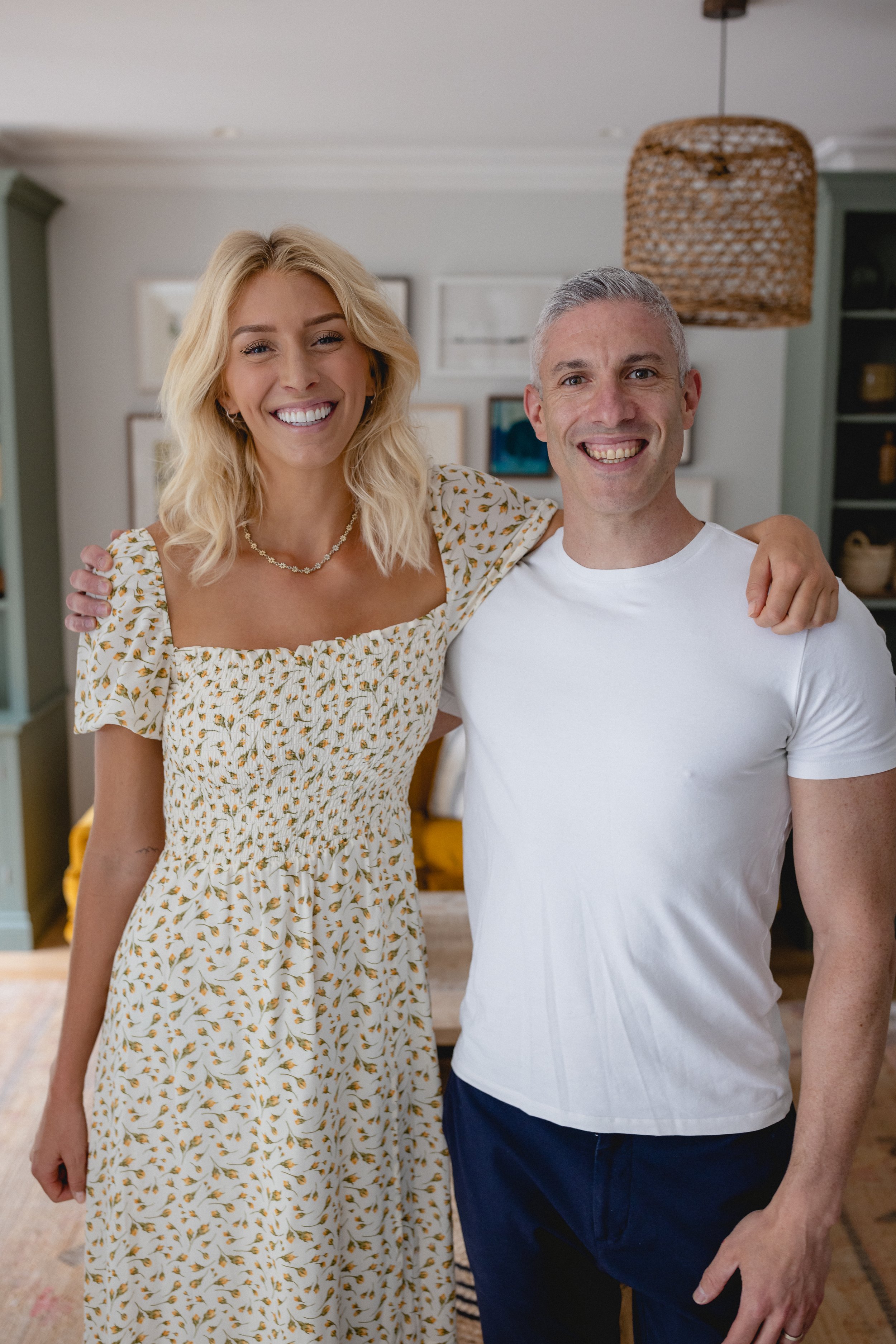 A smiling woman in a floral dress stands next to a smiling man in a white t-shirt. They are indoors, in a room with framed pictures on the wall and a wicker lampshade hanging from the ceiling.