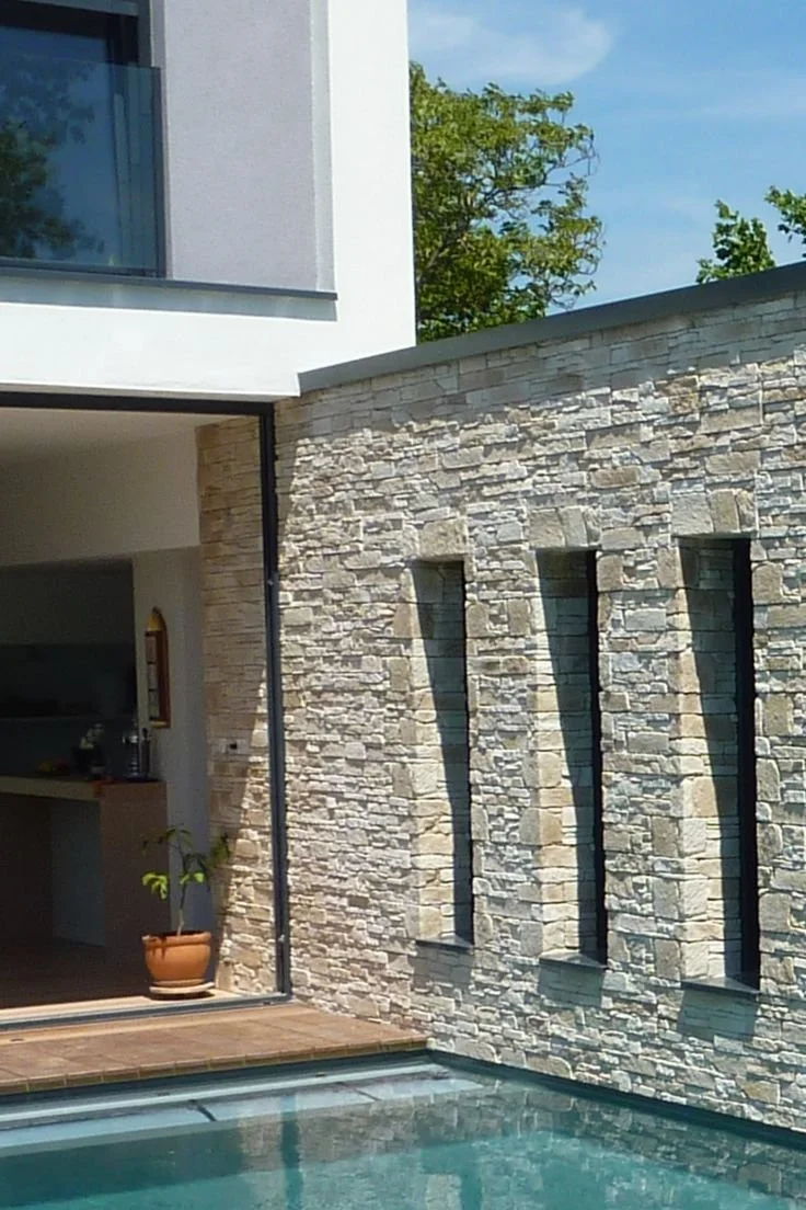 Une façade moderne avec un mur en pierre, trois fenêtres étroites et un coin de piscine, sous un ciel bleu, avec des arbres en arrière-plan.