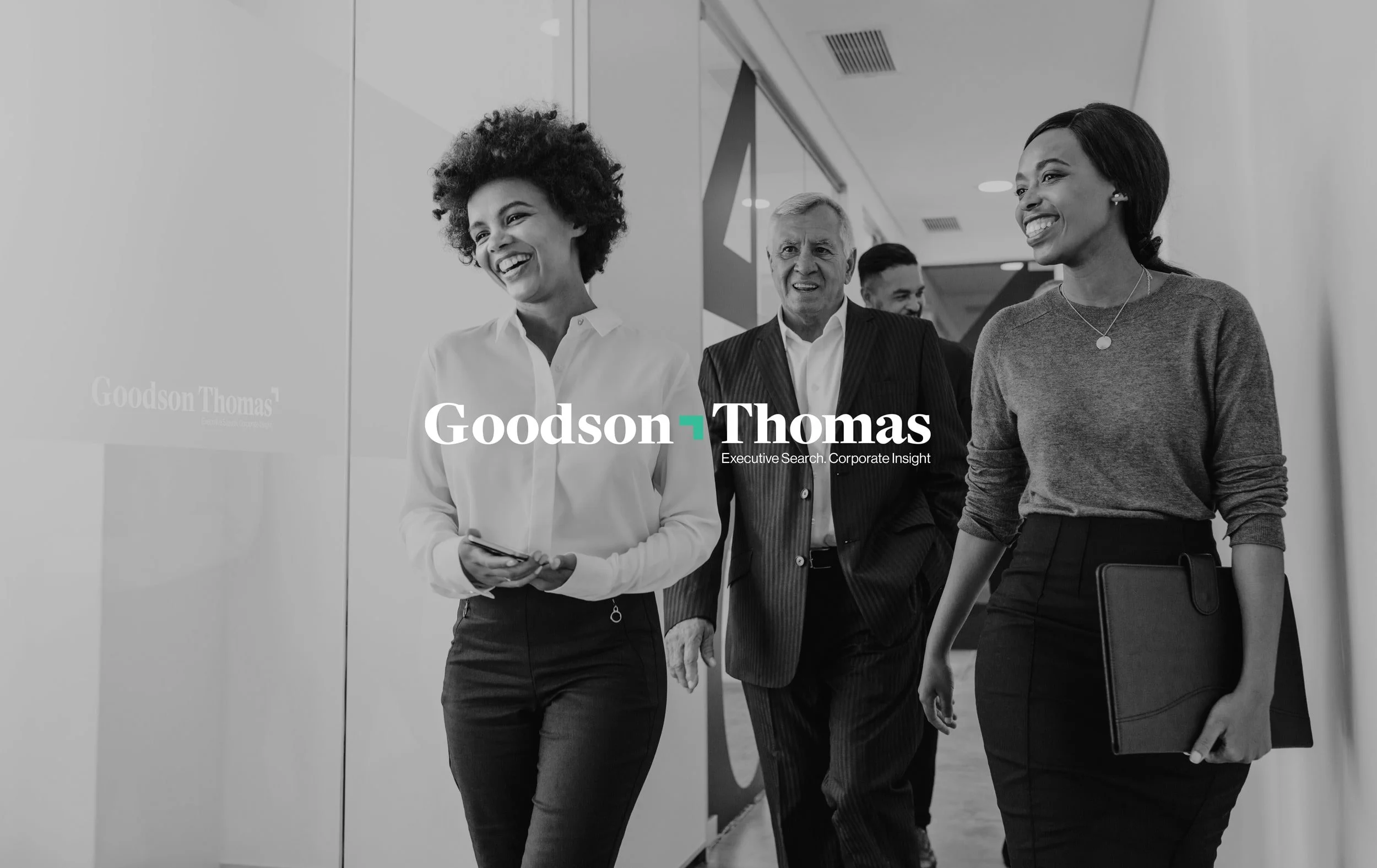 Group of business professionals walking and talking in an office hallway, two women smiling, one man in a suit. Black and white photo with 'Goodson Thomas' logo and tagline.