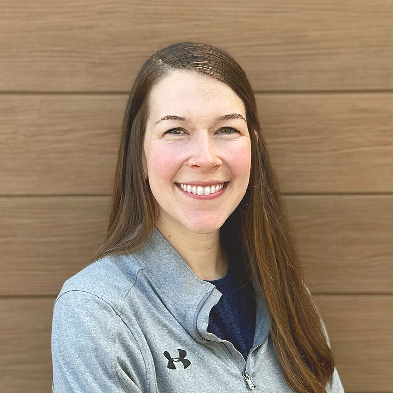 A woman with long brown hair smiling and crossing her arms, wearing a gray Under Armour zip-up jacket, standing in front of a wooden wall.