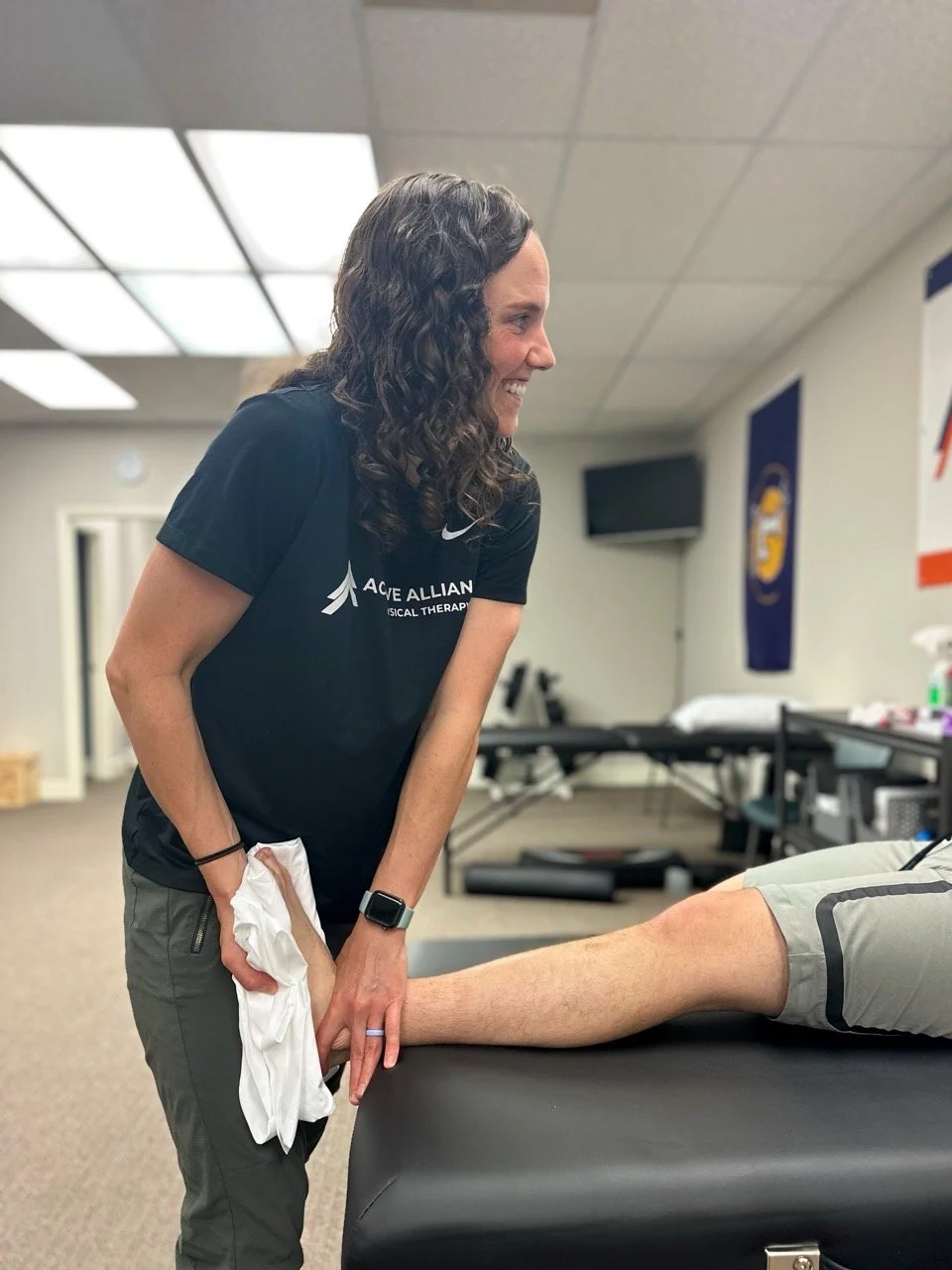 A physical therapist smiling while working on a patient's leg in a clinical setting.