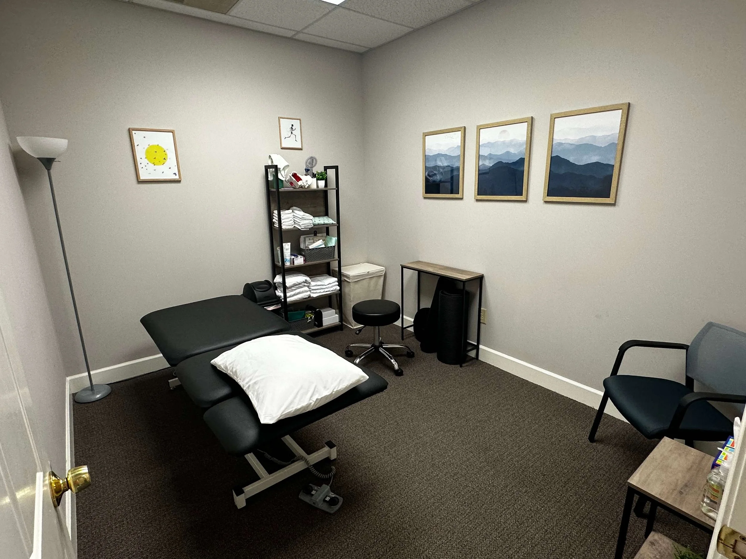 A small examination or therapy room with a black treatment table, a pillow, a chair, and a small shelf with folded towels. The walls have framed pictures of mountains and a running figure. There is a floor lamp in the corner.