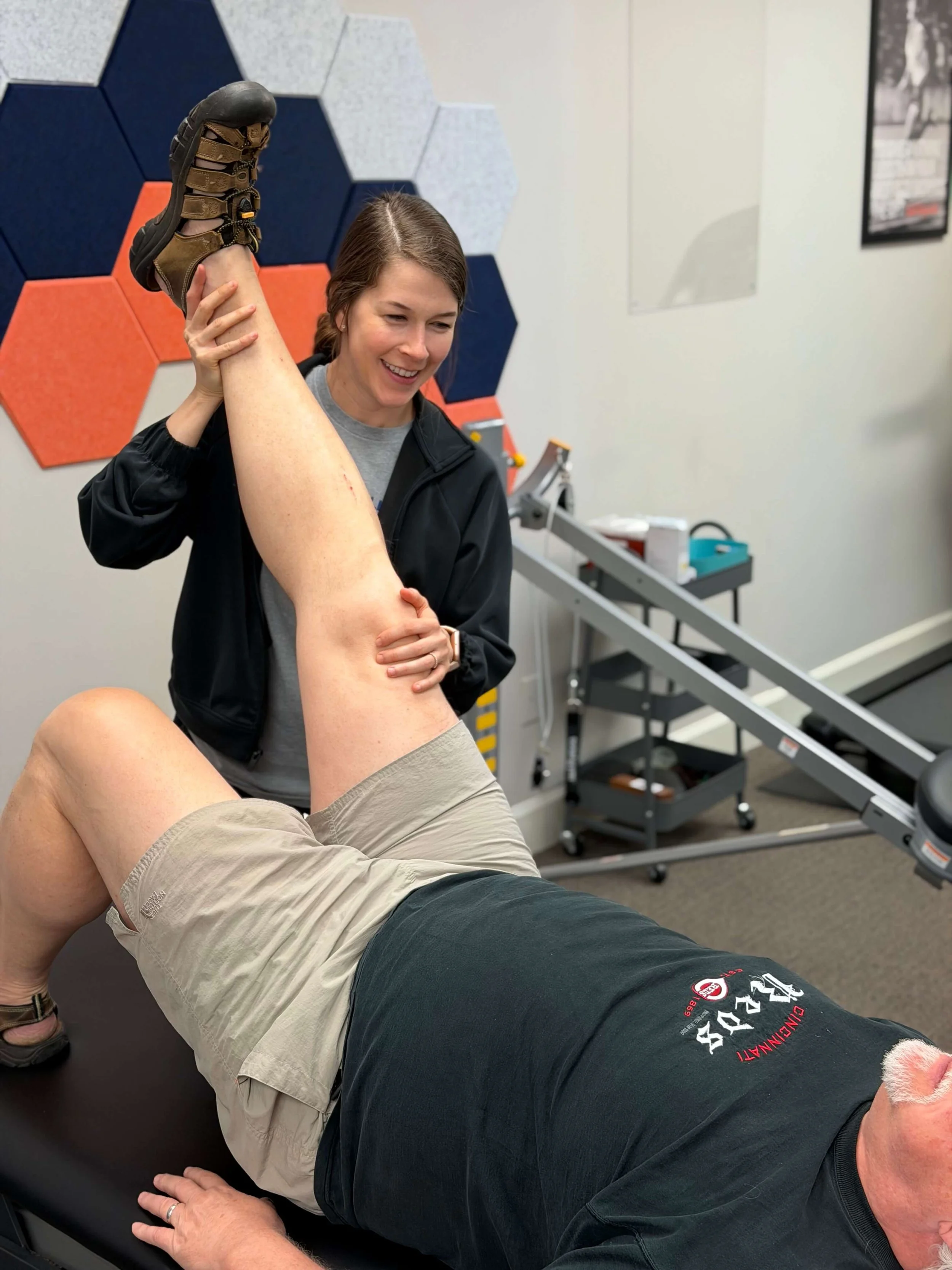 A woman assisting a man during physical therapy by supporting his leg while he lies on a therapy table.