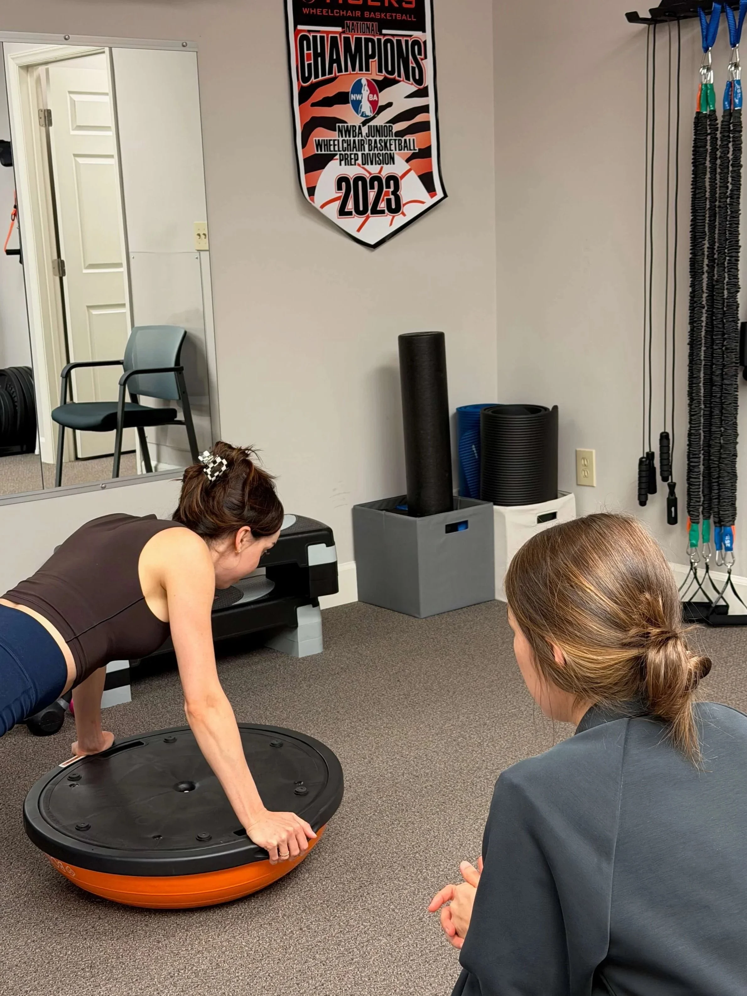 erforming a fitness exercise using a balance ball, with one woman in a plank position on the floor and another sitting nearby. The room has workout equipment against the wall, including resistance bands, foam rollers, and a banner celebrating a 2023 basketball championship.