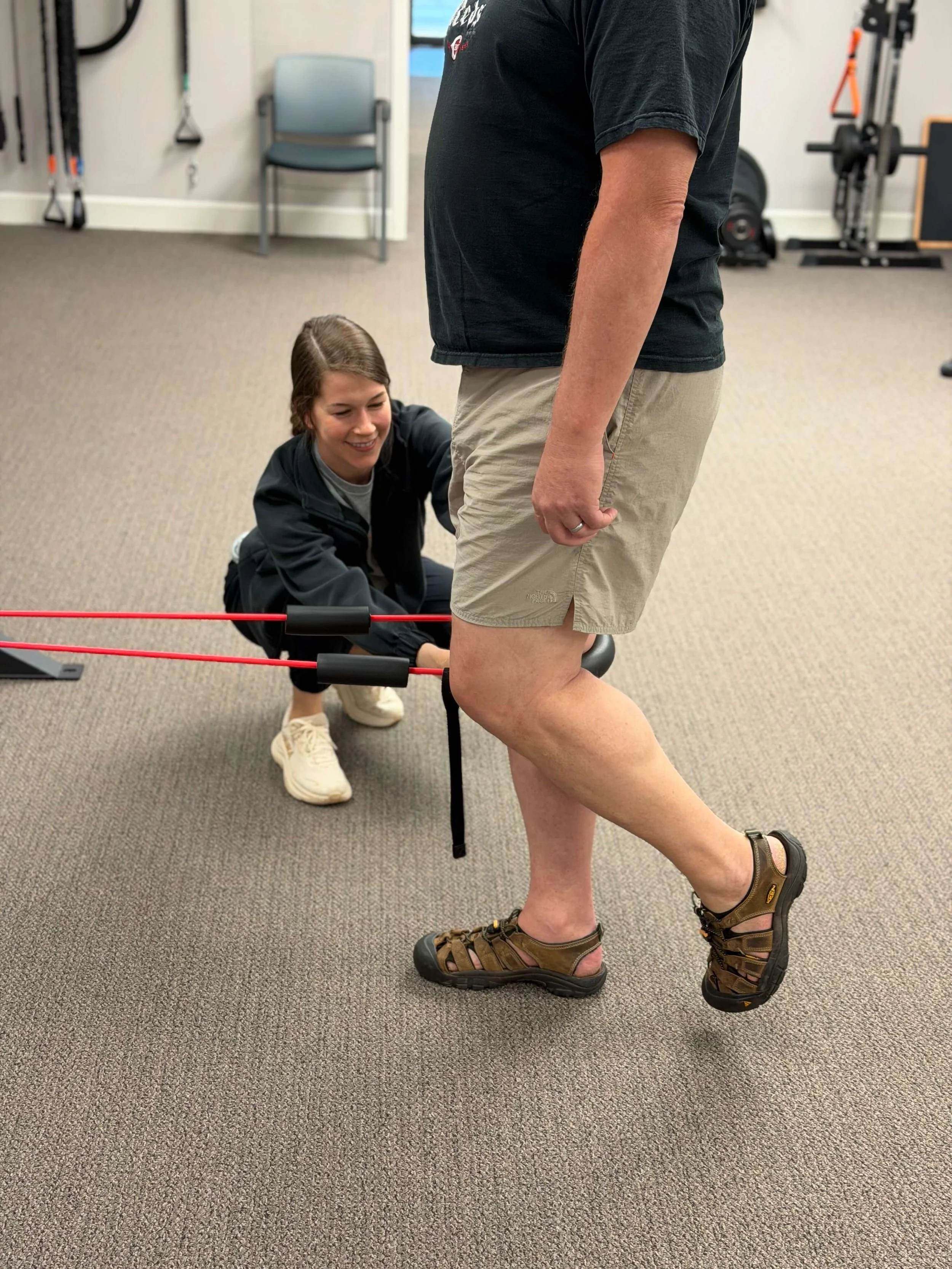 A woman sitting on the floor helping a man exercise his knee. The man is standing, wearing shorts and sandals. The woman is smiling, wearing a black jacket, and white sneakers. The room has gym equipment in the background.