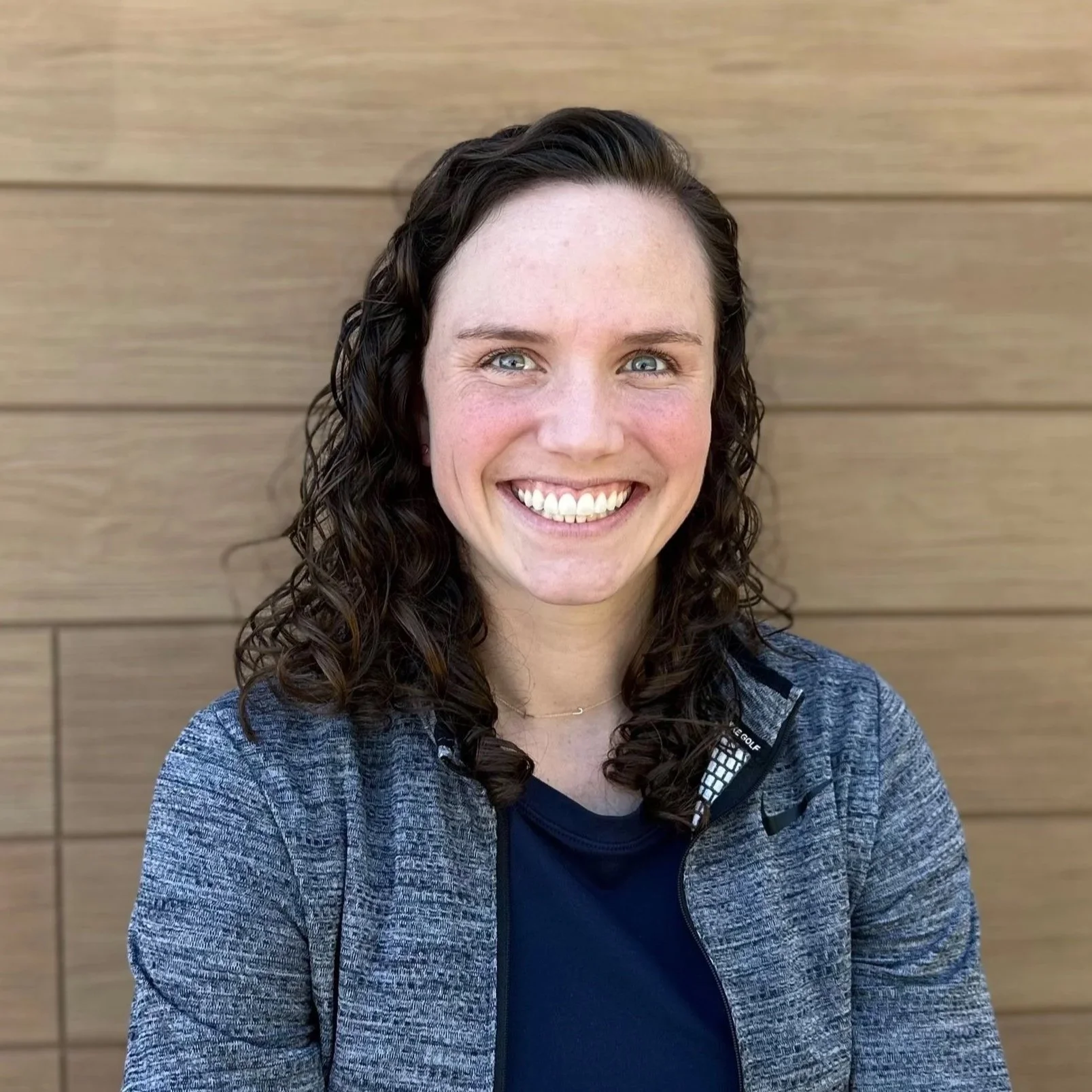 A person with curly hair smiling and crossing their arms, standing against a wooden wall.