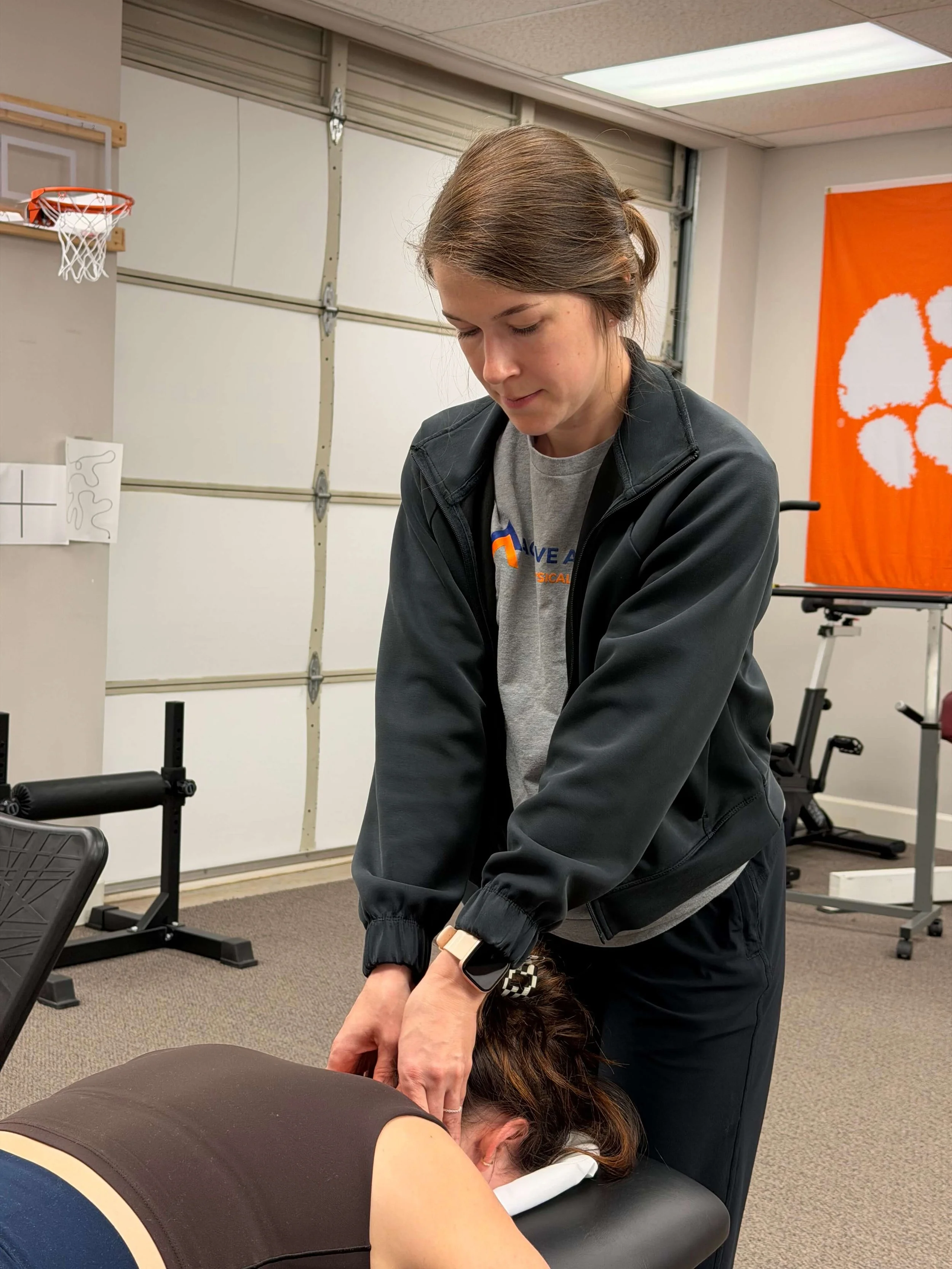 A woman receiving cervical manual therapy, with a person lying face down on a massage table, in a clinic with an orange Clemson University banner in the background.