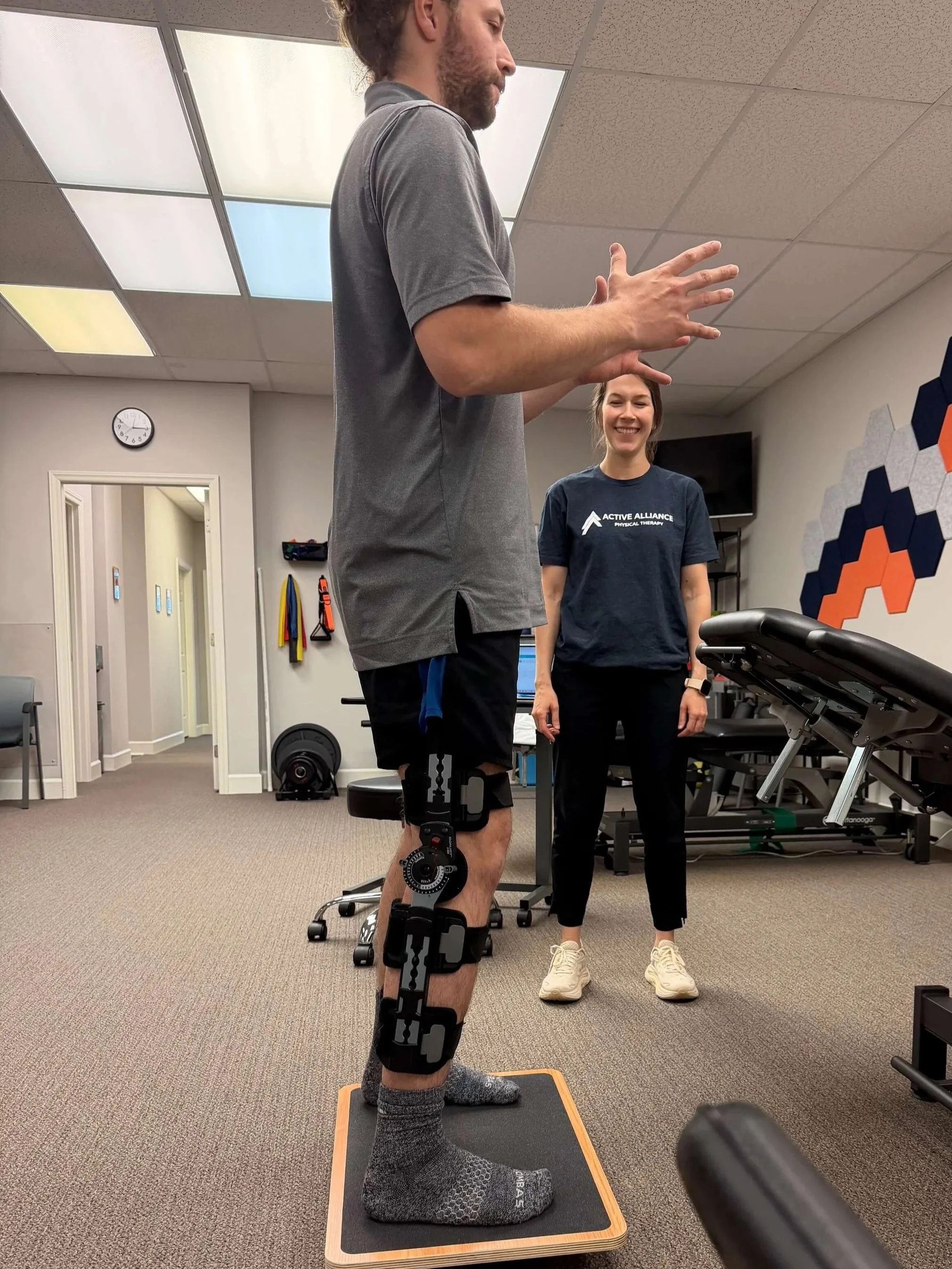 A man is standing on a balance board with a post surgical brace on his leg, while a woman in looks on and smiles in a physical therapy or rehabilitation setting.