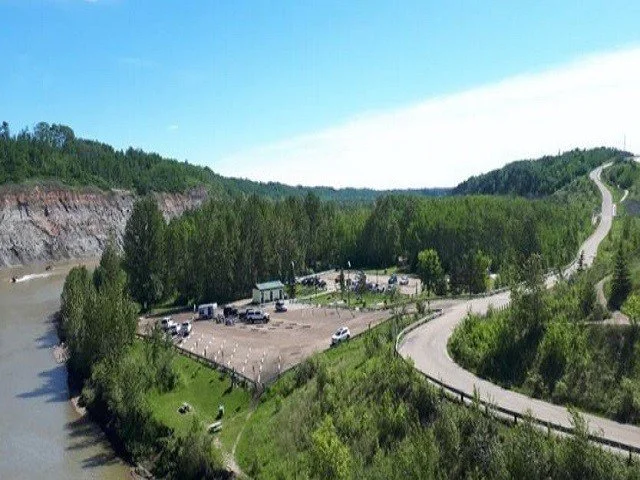 A scenic view of a river, surrounded by green trees, a parking lot with cars, and a winding road through a hilly landscape at a sunny day.