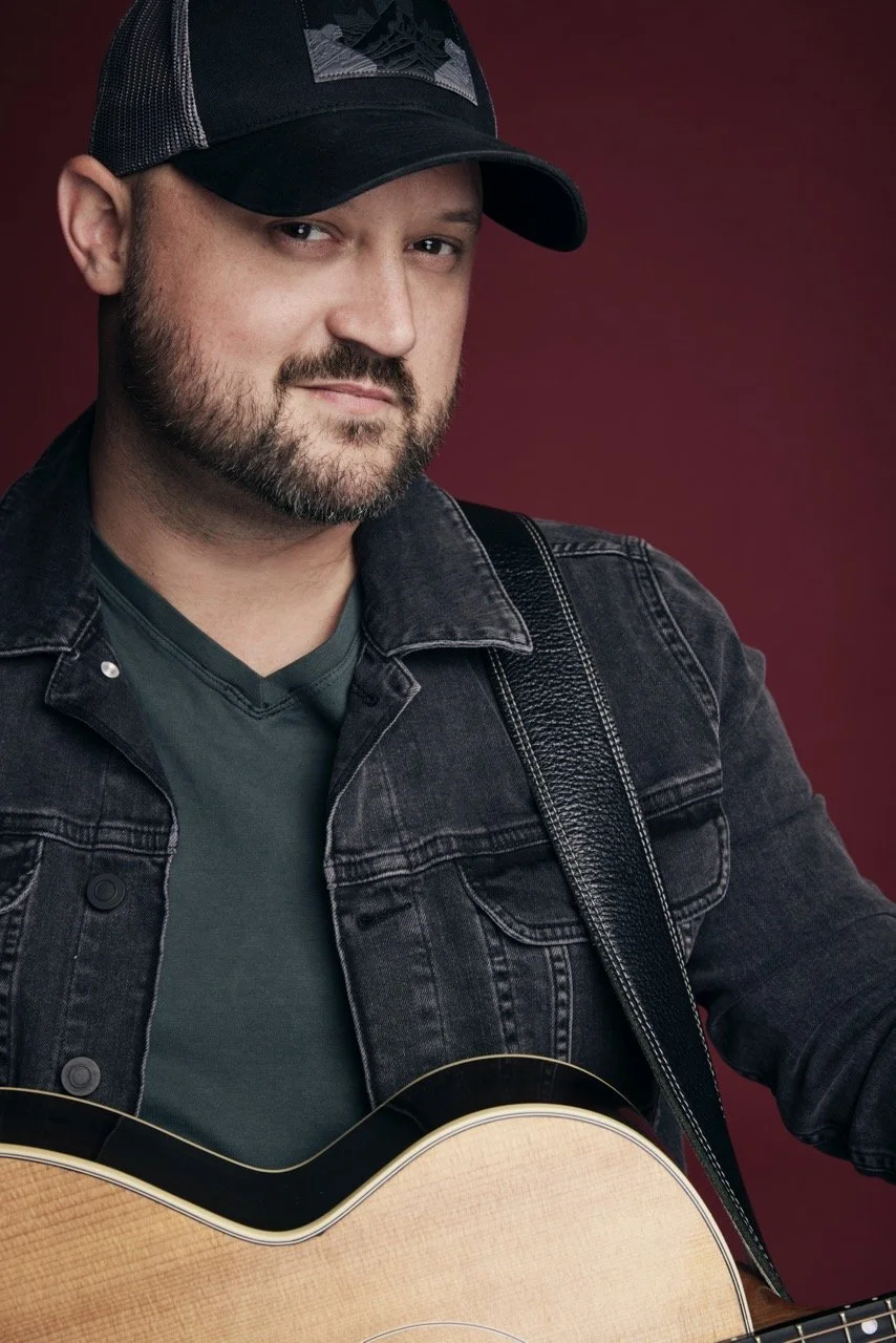 A man with a beard and mustache, wearing a black baseball cap, black denim jacket, and dark green shirt, holding a light-colored acoustic guitar, posing against a red background.