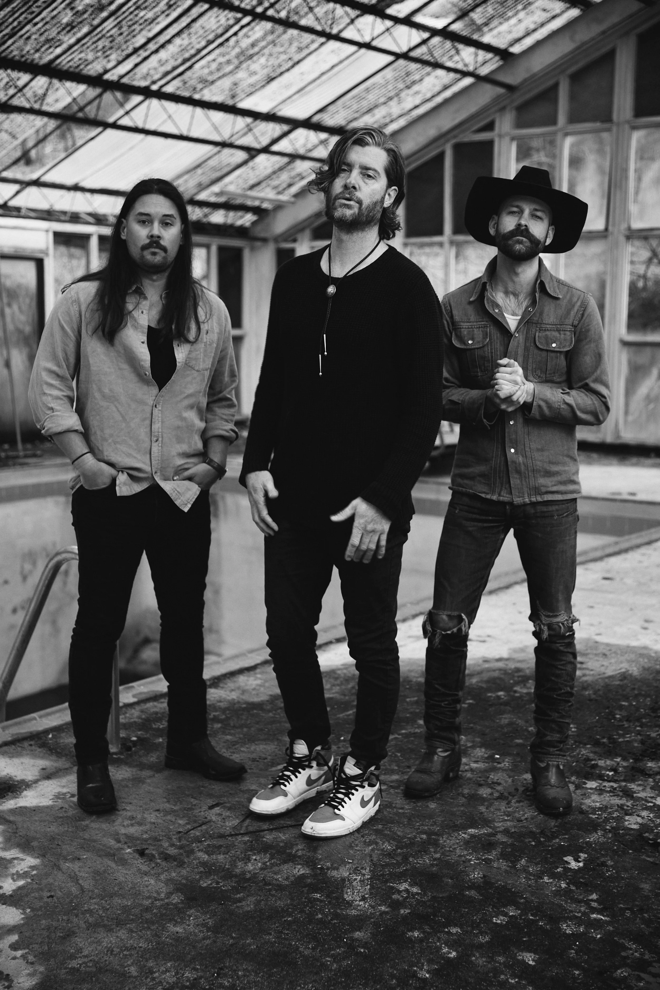 Black and white photo of three men standing inside a greenhouse, with the man in the middle wearing a black sweater and sneakers, the man on the right wearing a wide-brimmed hat and denim jacket, and the man on the left in a button-up shirt.