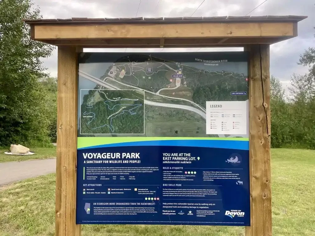 Information board at Voyageur Park with a map, legend, and rules about the park's trails, wildlife, and activities. The sign is mounted on a wooden frame outdoors with trees and grass in the background.