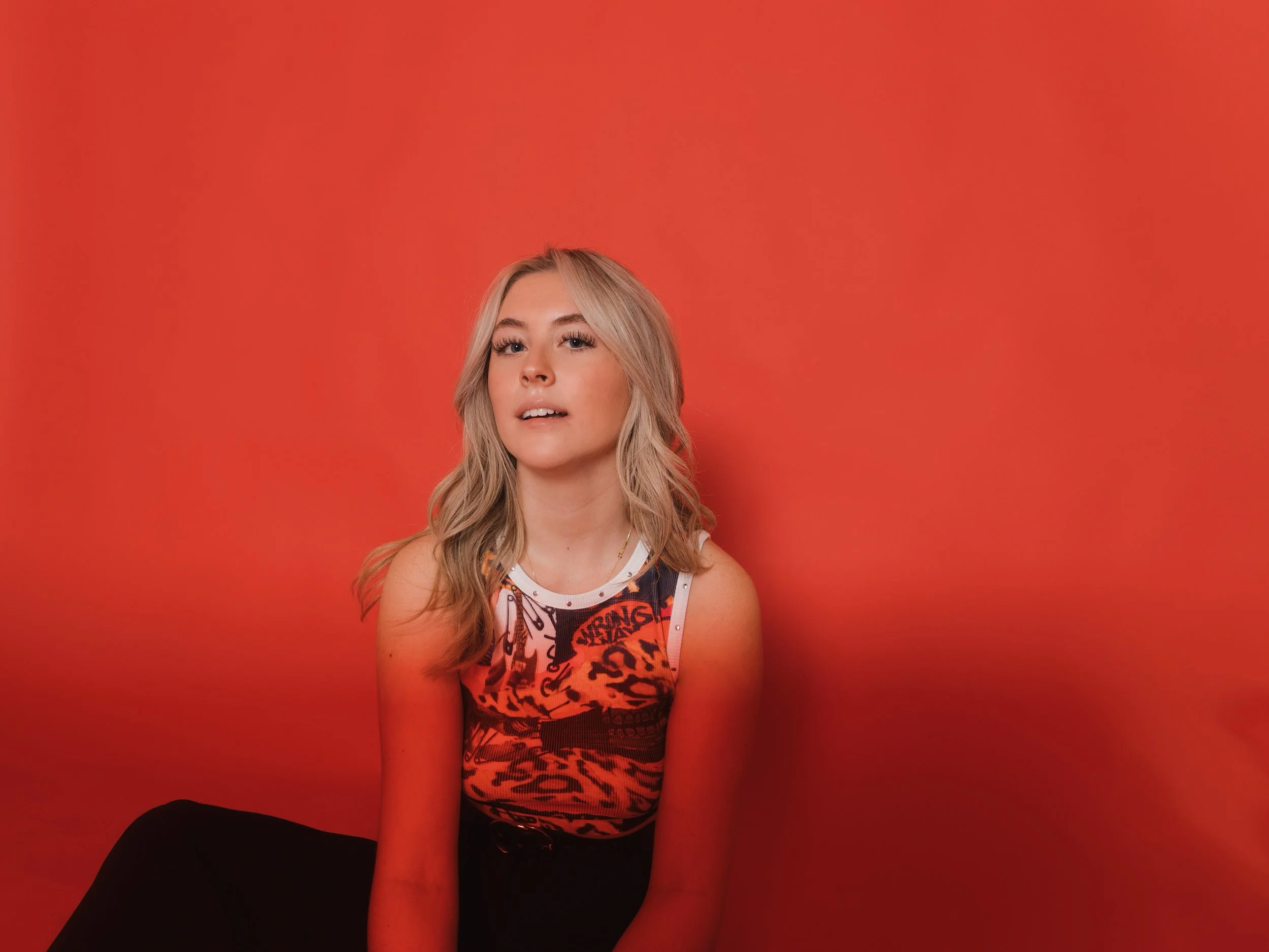 A young woman with blonde hair sitting in front of a red background, wearing a tank top with a red and black pattern.
