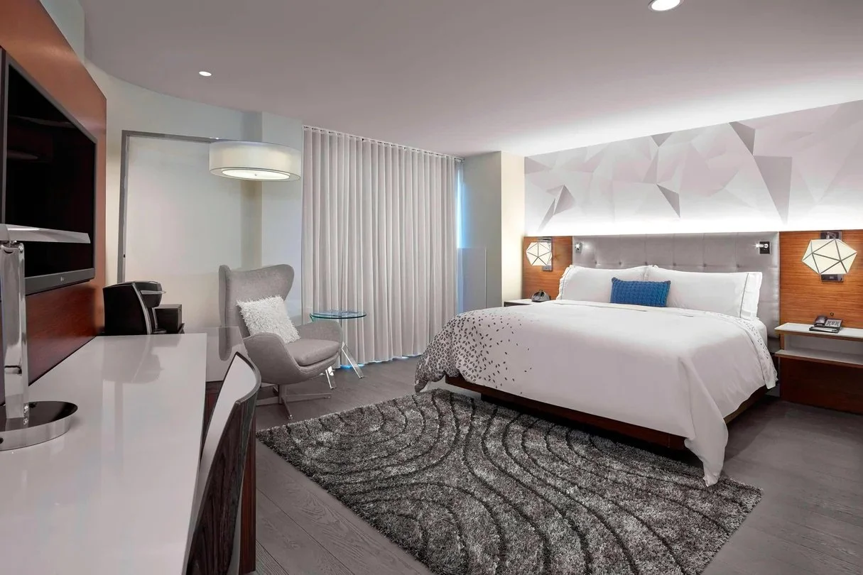Modern hotel room with a large bed, grey headboard, blue accent pillow, bedside lamps, and a textured white wall. There is a sitting area with a grey armchair, a side table, and a window with white curtains. A white desk with a television and other furniture is also visible.