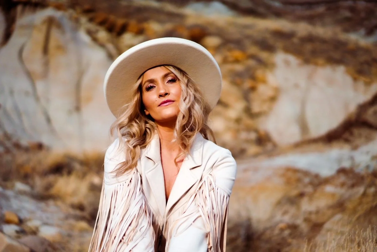 A woman with blonde, wavy hair wearing a wide-brimmed hat and a white fringed jacket outdoors in a rocky landscape.