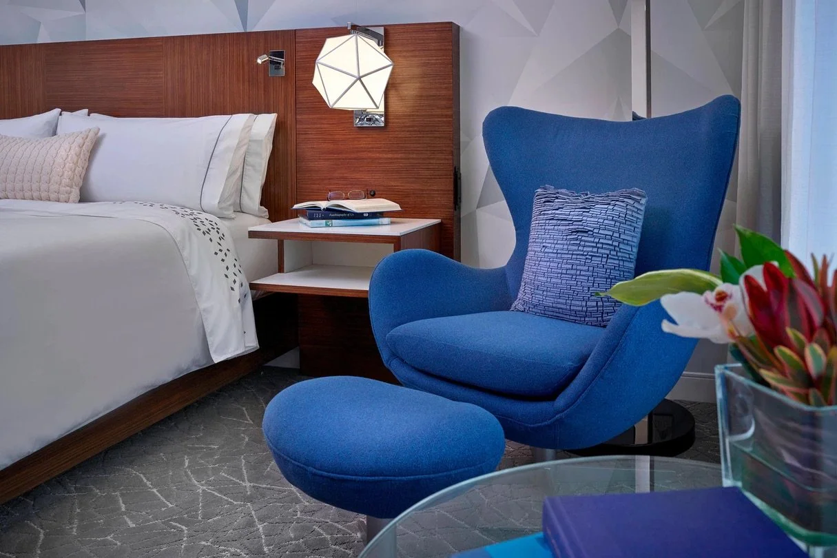 A bedroom featuring a white bed with pillows, a wooden headboard, a blue armchair with a patterned cushion, a nightstand with books and glasses, and a flower arrangement on a glass table.