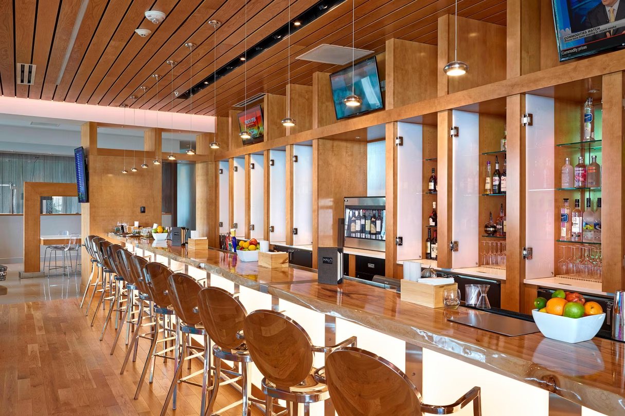 Empty bar with wooden counter, bar stools, and shelves with liquor bottles and glassware.