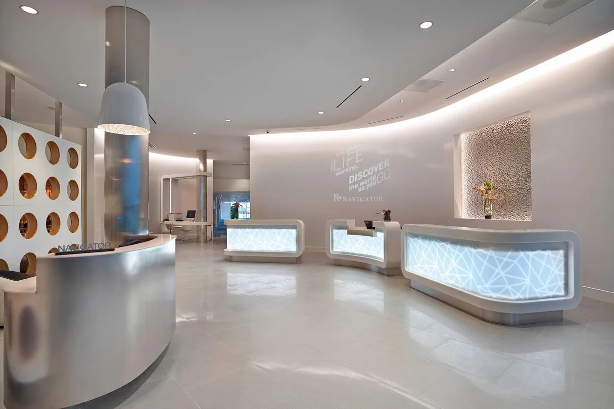 Modern hospital or clinic reception area with white illuminated counters, a curved front desk, and a wall with a projection that says 'LIFE working DISCOVER the world as you GO'. The space has a sleek, minimalist design with soft lighting and a neutral color palette.