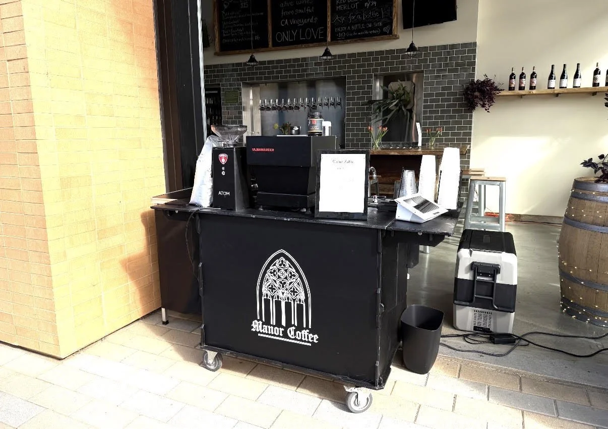 Coffee stand with Manor Coffee logo, espresso machine, paper cups, and a cooler inside a cafe. Shelves with bottles and plants in the background.
