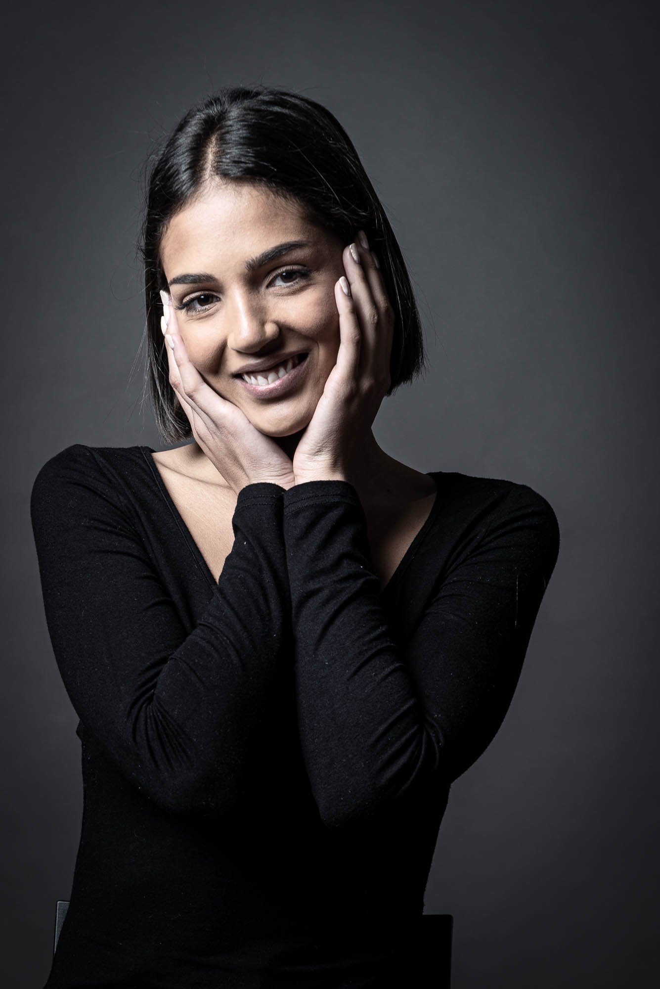Mujer sonriente con cabello oscuro y corto, vistiendo camisa negra, sostiene su rostro con las manos.